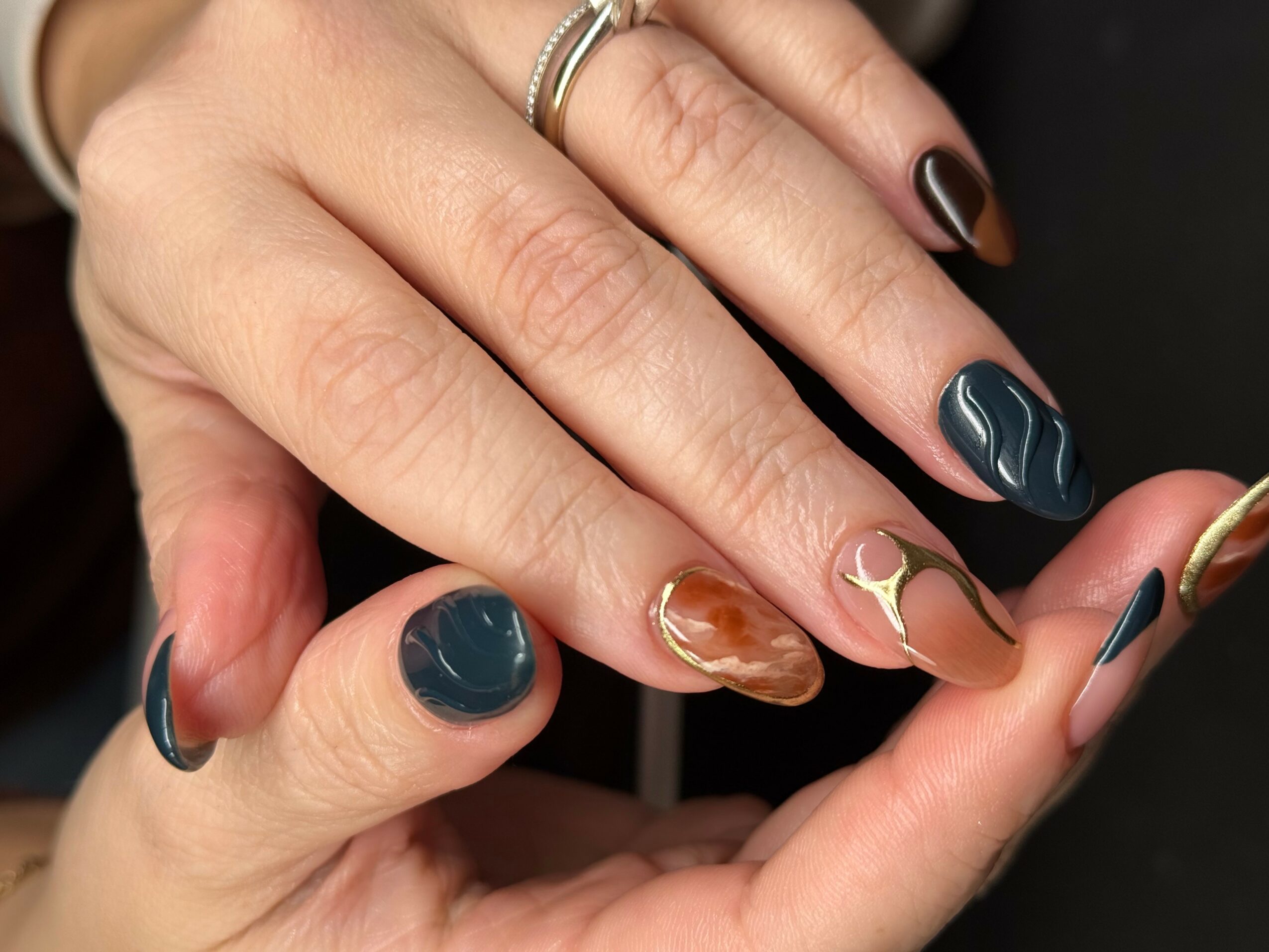 Close-up of hands with manicured nails in dark blue, brown, and beige with gold accents, and rings on fingers.