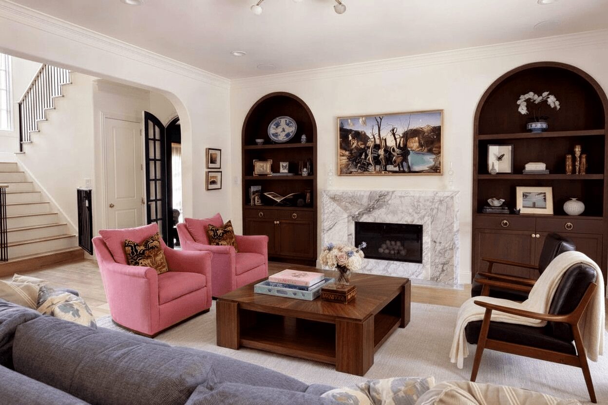 Living room with pink armchairs, a wooden coffee table, a fireplace, and built-in dark wood shelves, with a staircase visible on the left.