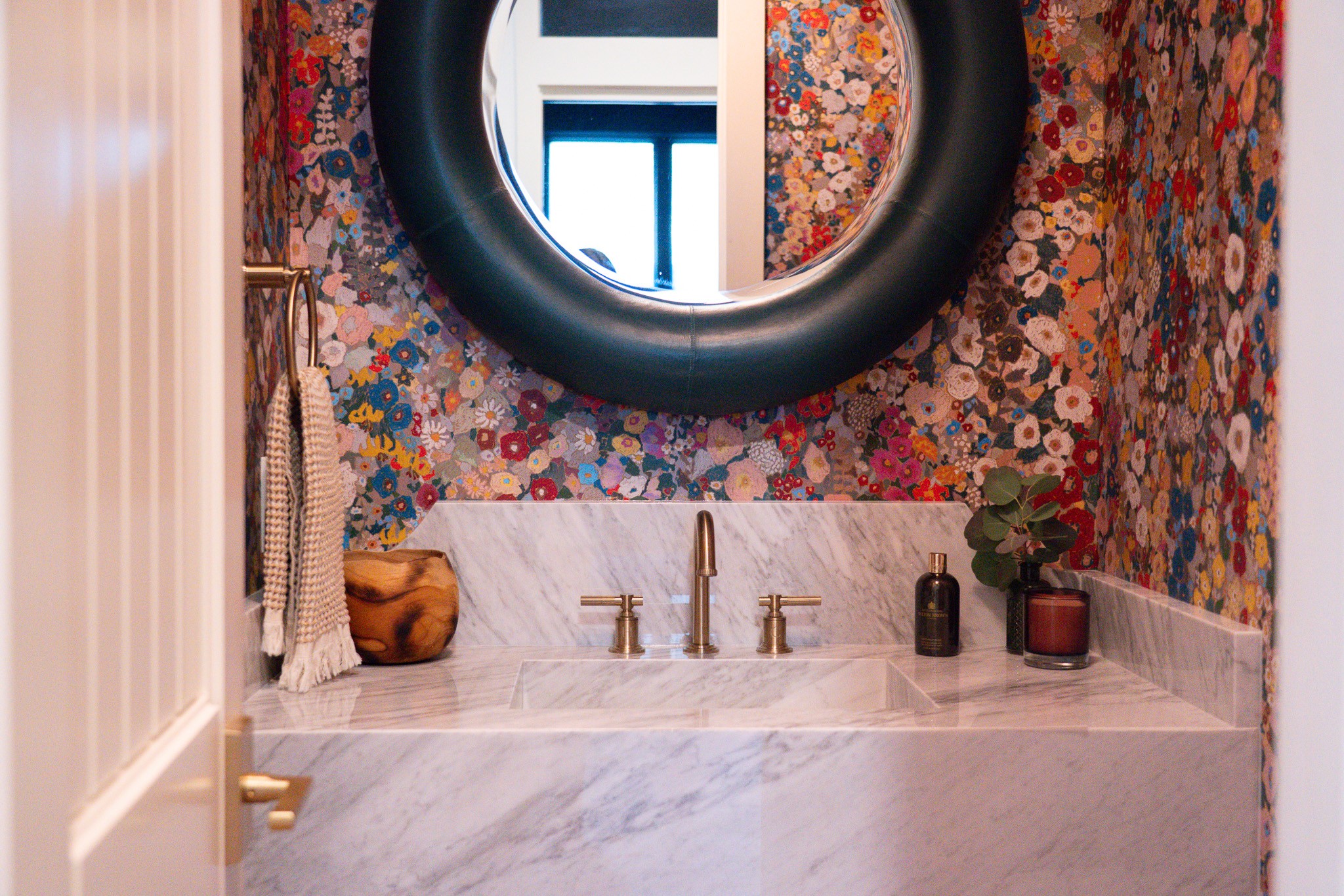 Bathroom sink with a round mirror above, colorful floral wallpaper, and a marble countertop with various items.