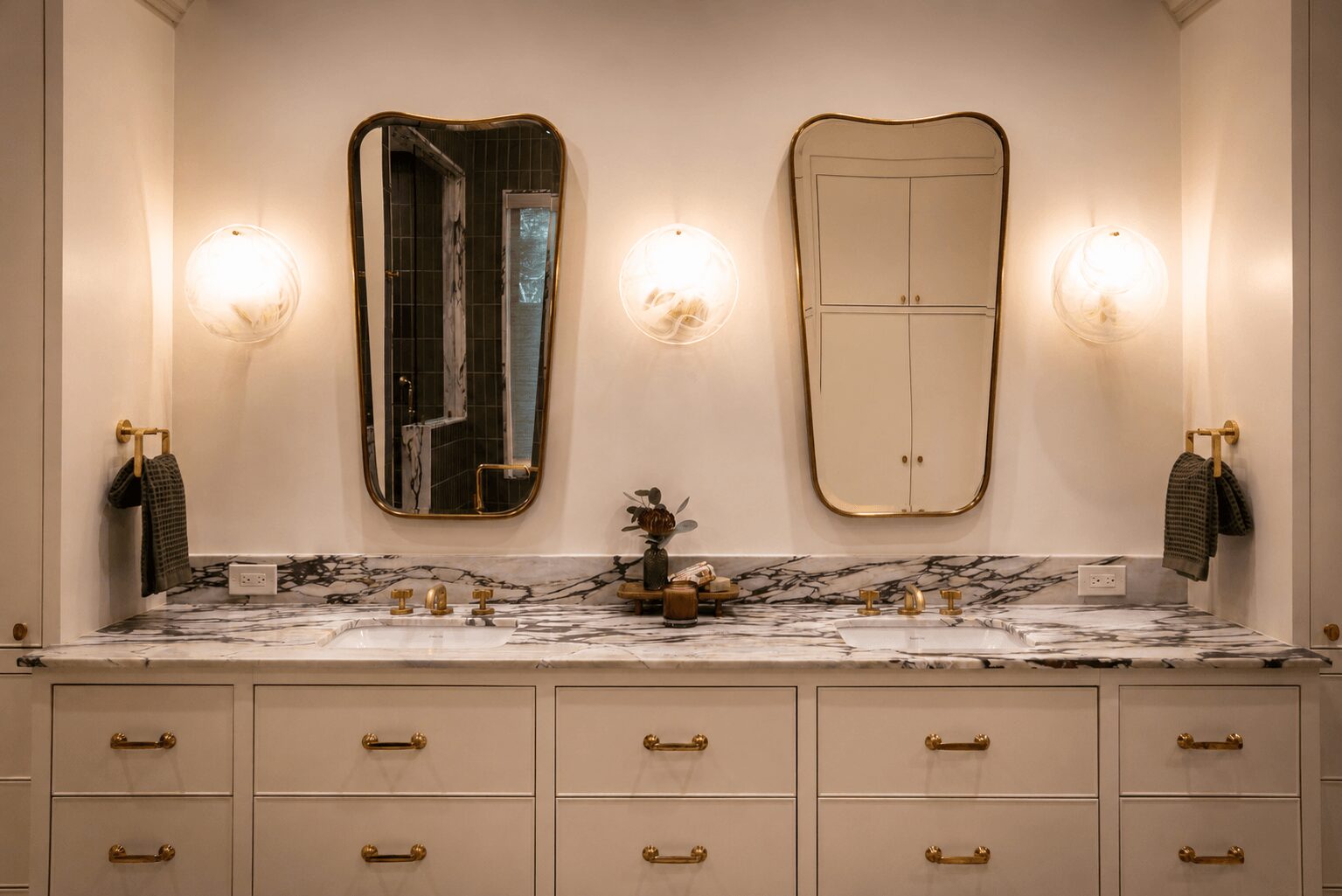 Bathroom vanity with two mirrors, two wall sconces, and a marble countertop with toiletries, against a white wall.