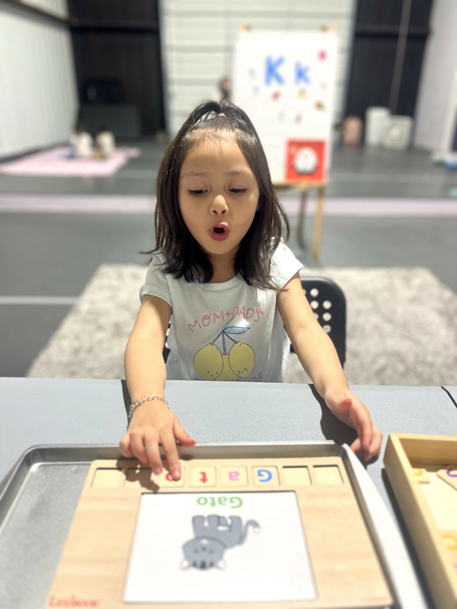 Young girl with shoulder-length hair looks surprised at a table with educational materials, in a classroom setting.