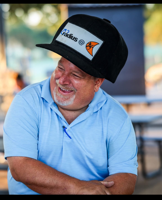 Smiling man wearing a black cap and light blue polo shirt outdoors, with blurred background.