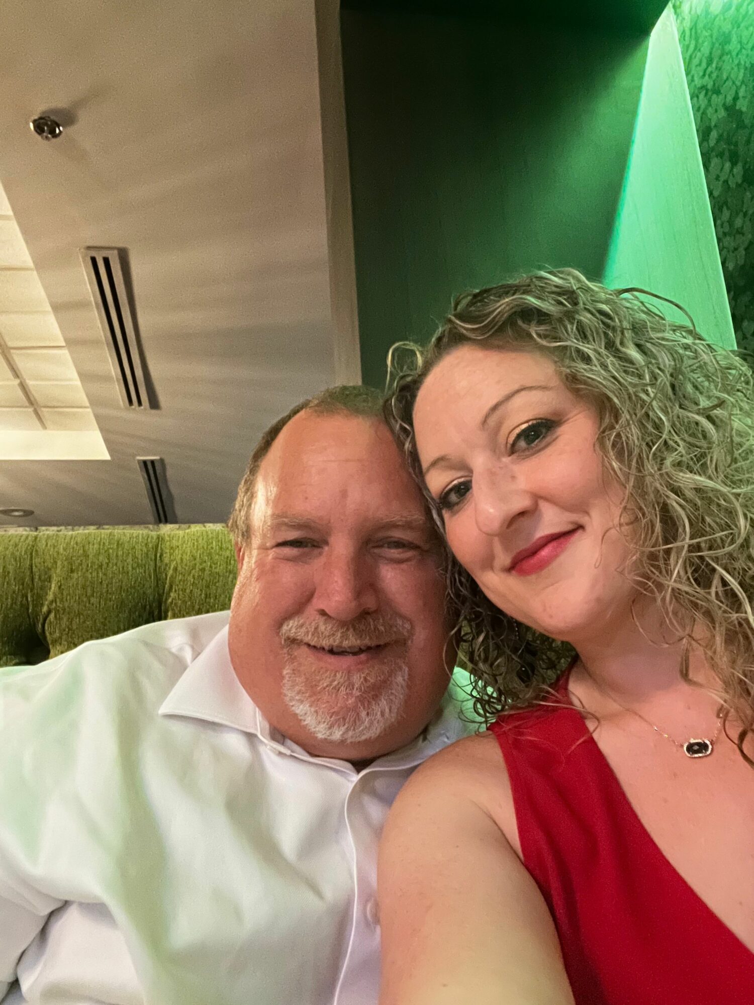 A woman with curly hair and a man with a beard and mustache are close together, smiling, indoors with a green background.