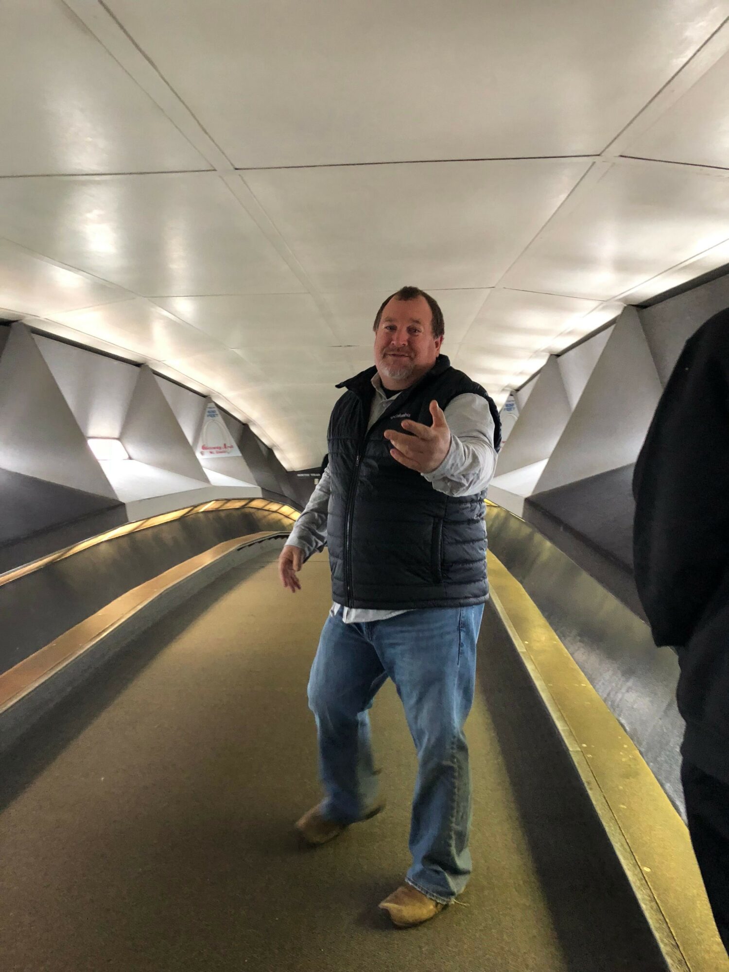 Person standing on a curved yellow line in an indoor corridor, giving a thumbs-up gesture, with white tiled wall behind.