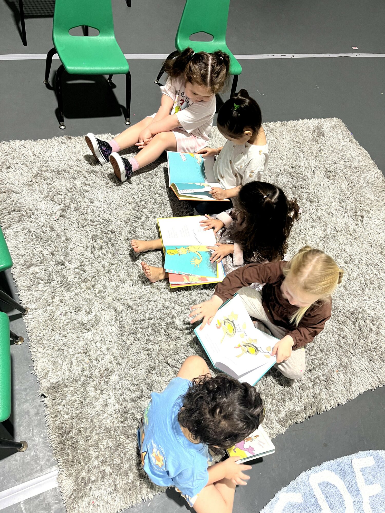Four children sitting on a gray rug, reading colorful books, with green chairs around them.