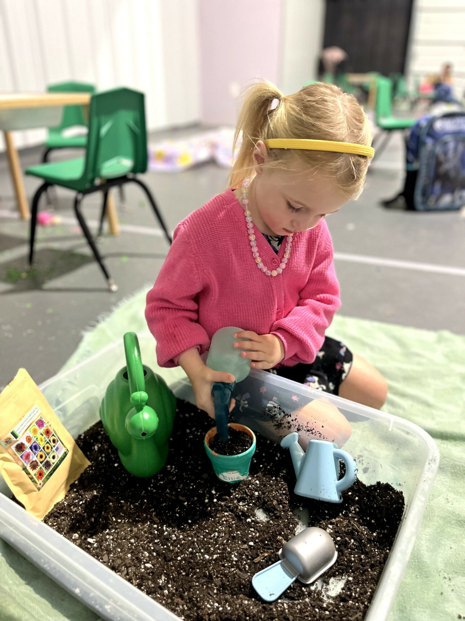 Young girl with blonde hair in a ponytail, wearing a pink sweater and pearl necklace, playing with gardening tools in a plastic container filled with soil.