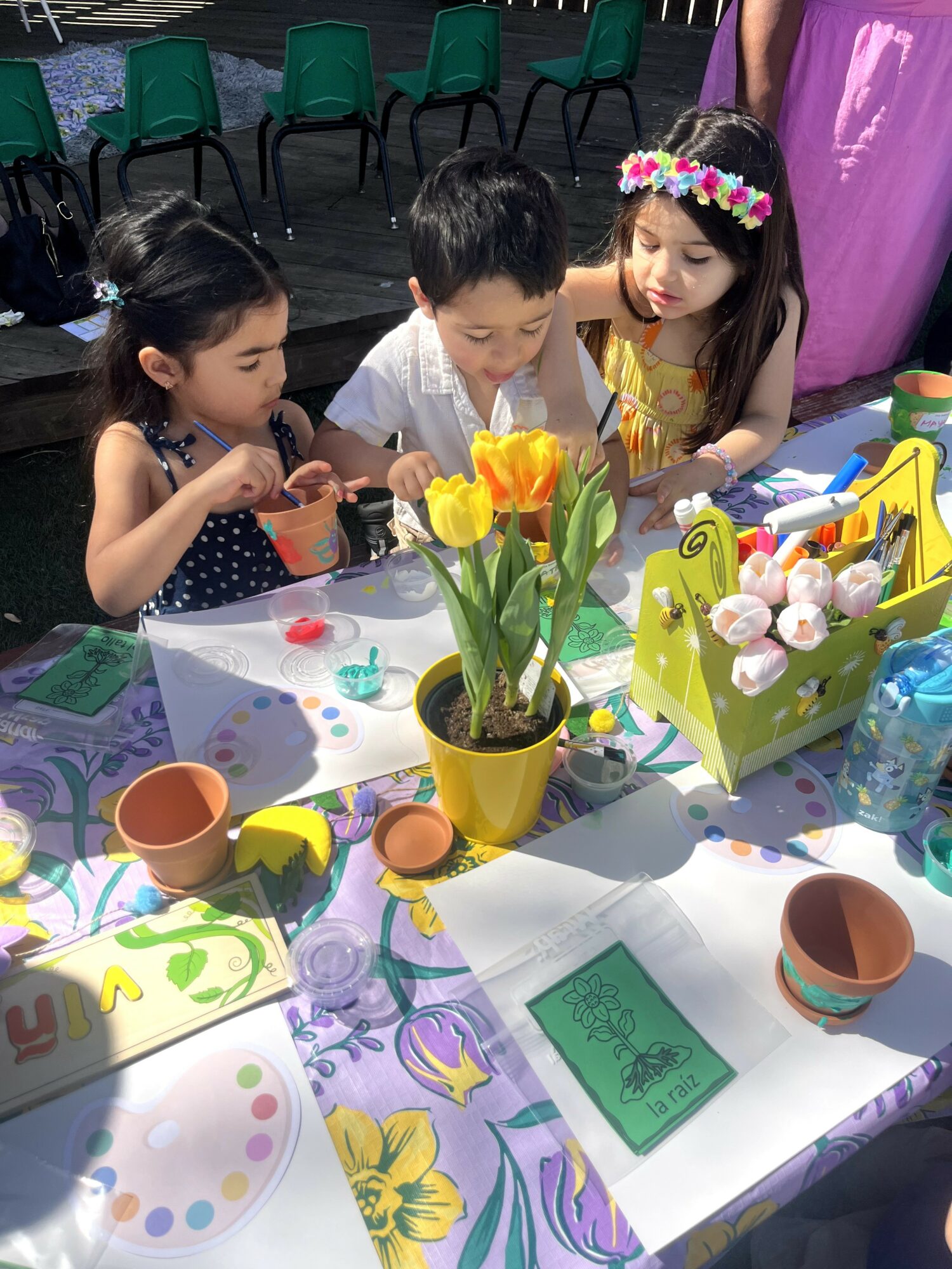 Three children sit at a table with colorful art supplies, flowers, and decorated eggs, engaging in a craft activity outdoors.