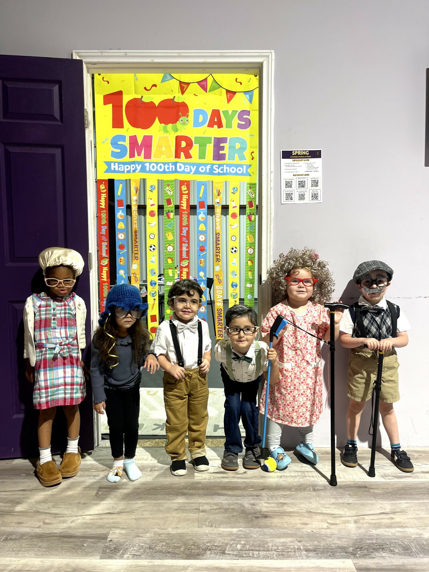 Six children stand in front of a colorful poster celebrating 100 days of school, smiling and holding hands.
