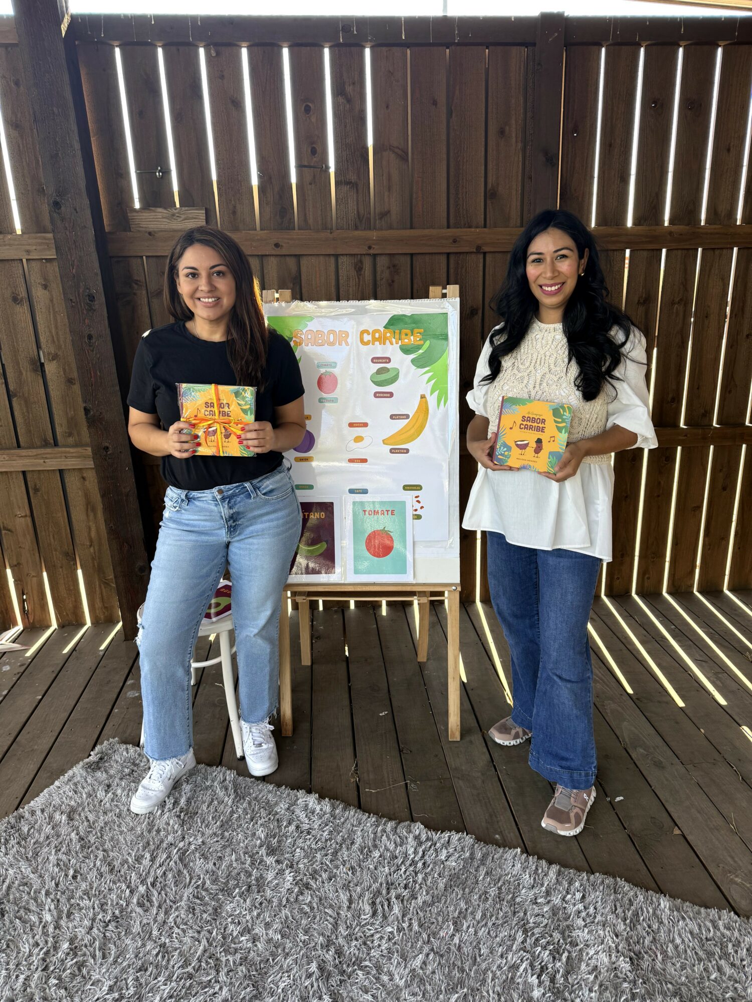 Two women stand outdoors in front of a wooden fence, holding books and smiling, with a poster on an easel between them.