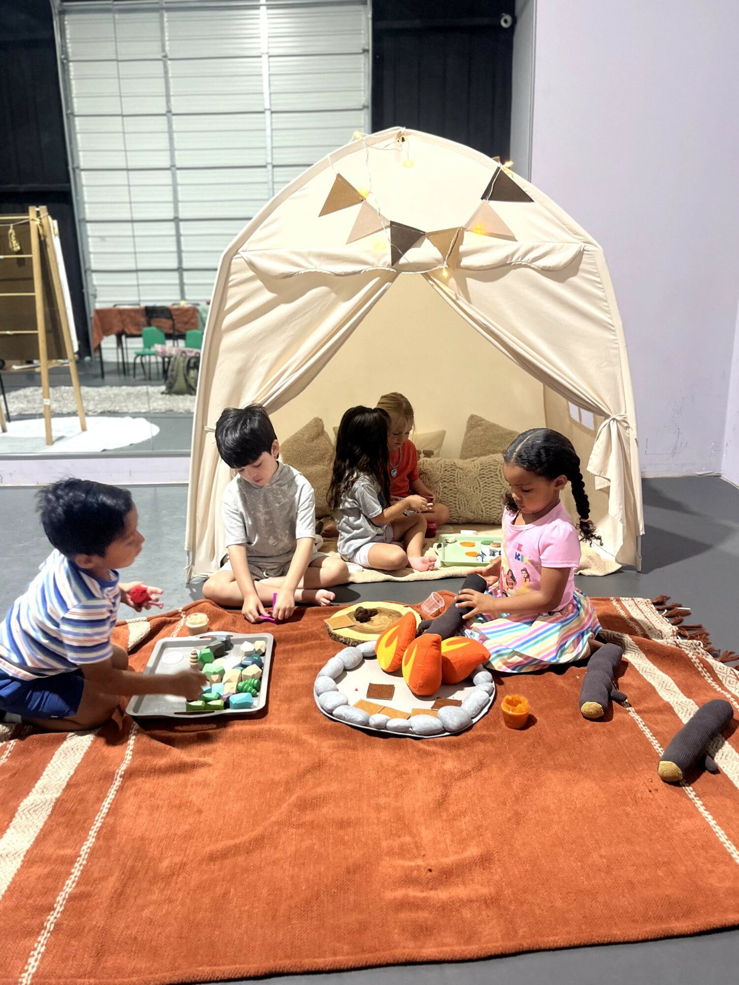 Children playing with toys inside a beige tent on a brown rug, with stuffed animals and toys around them.