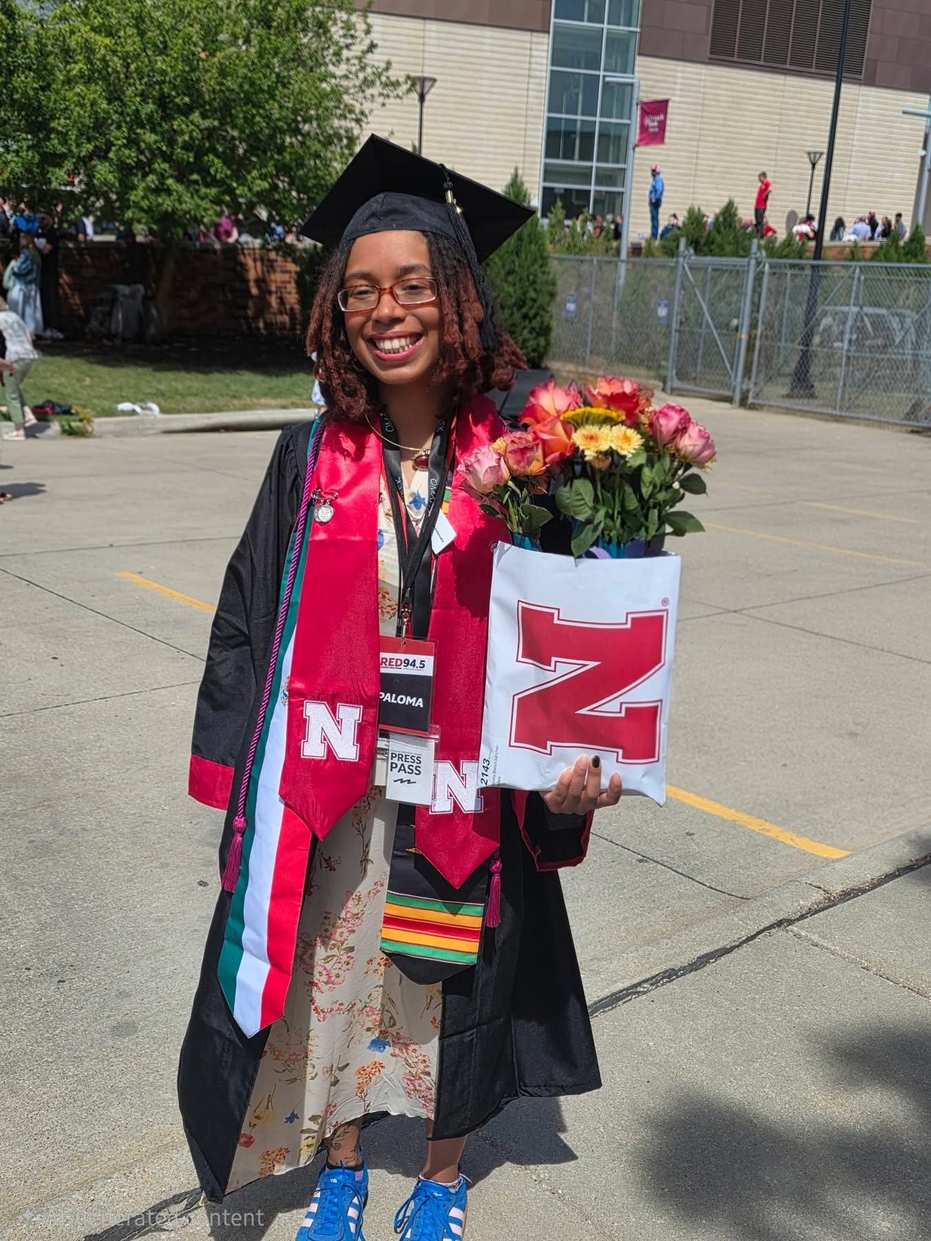 Young woman in graduation gown and cap holding a bouquet of flowers, smiling outdoors on a sunny day.