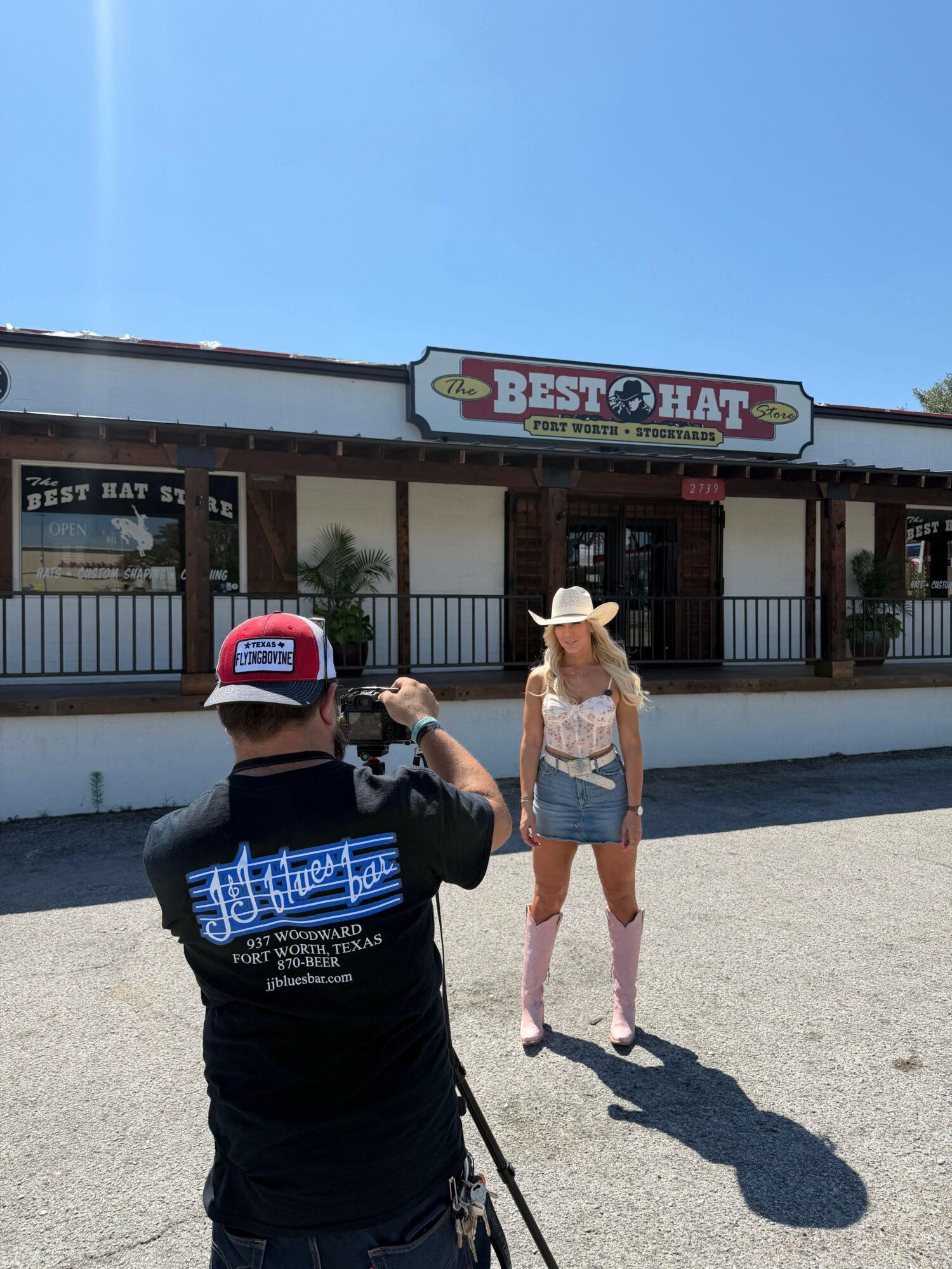 Person taking a photo of a woman in front of a store with a sign reading 'Best O'Rite' on a sunny day.
