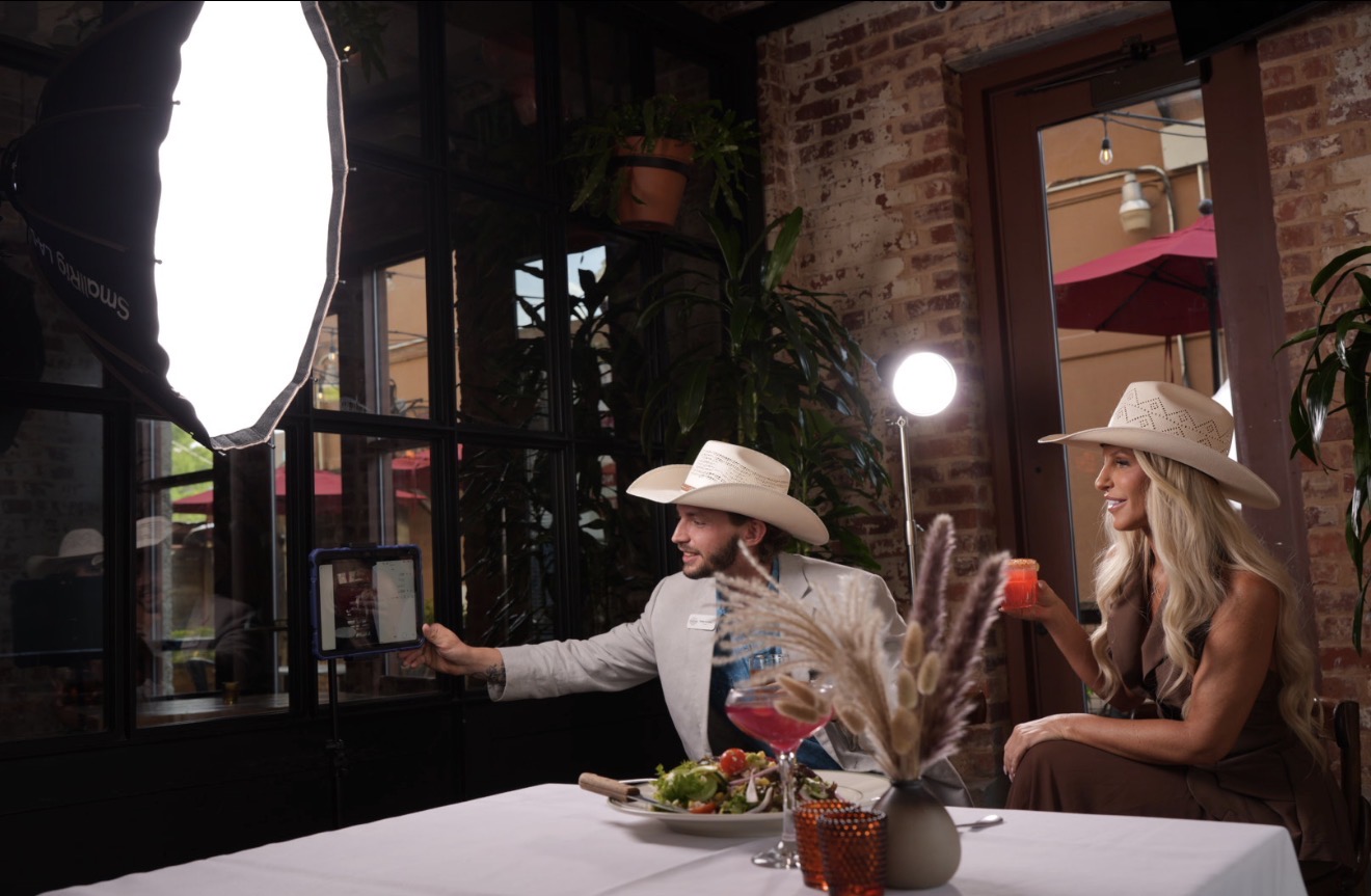 Two people wearing cowboy hats sit at a table with a white tablecloth, in a room with brick walls and plants.