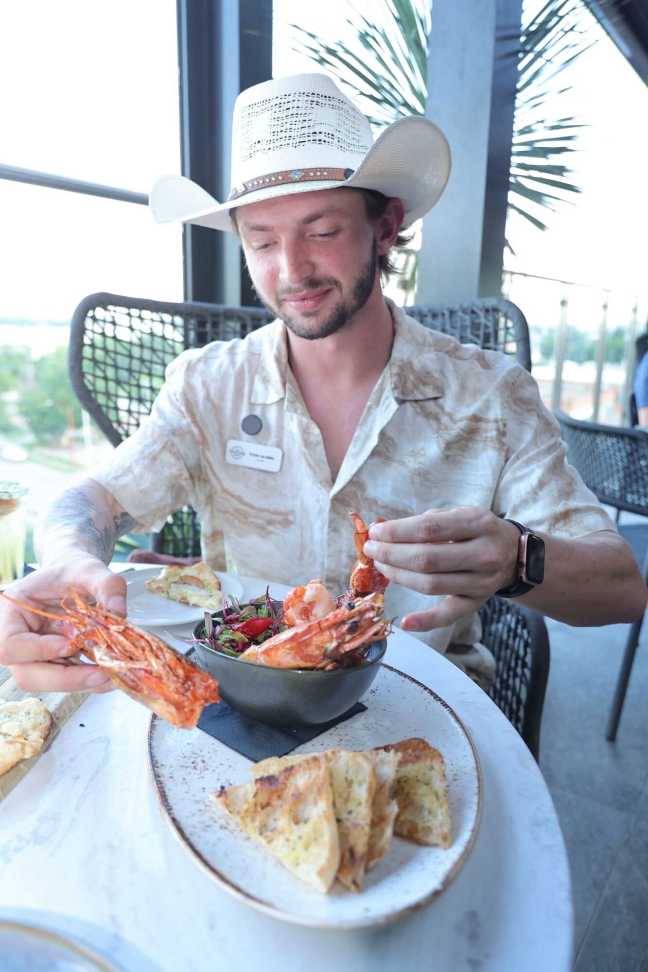 Person wearing a cowboy hat and patterned shirt holding a lobster claw with food on a plate, sitting at a table near a window.