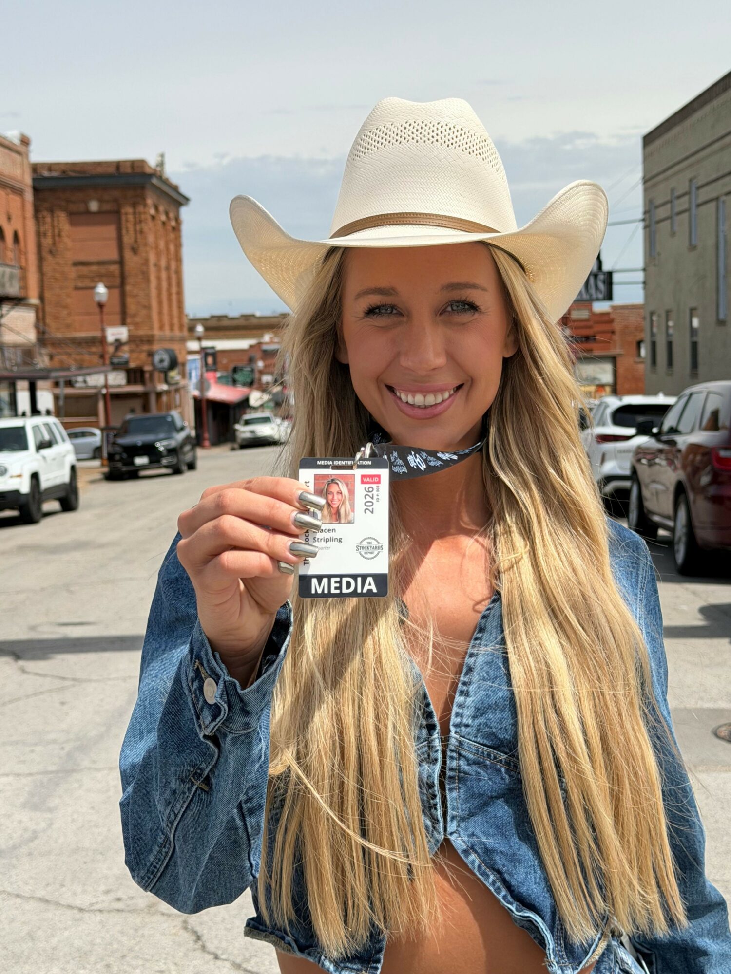 Young woman with long blonde hair, wearing a white hat and denim jacket, smiling and holding a media pass, outdoors.
