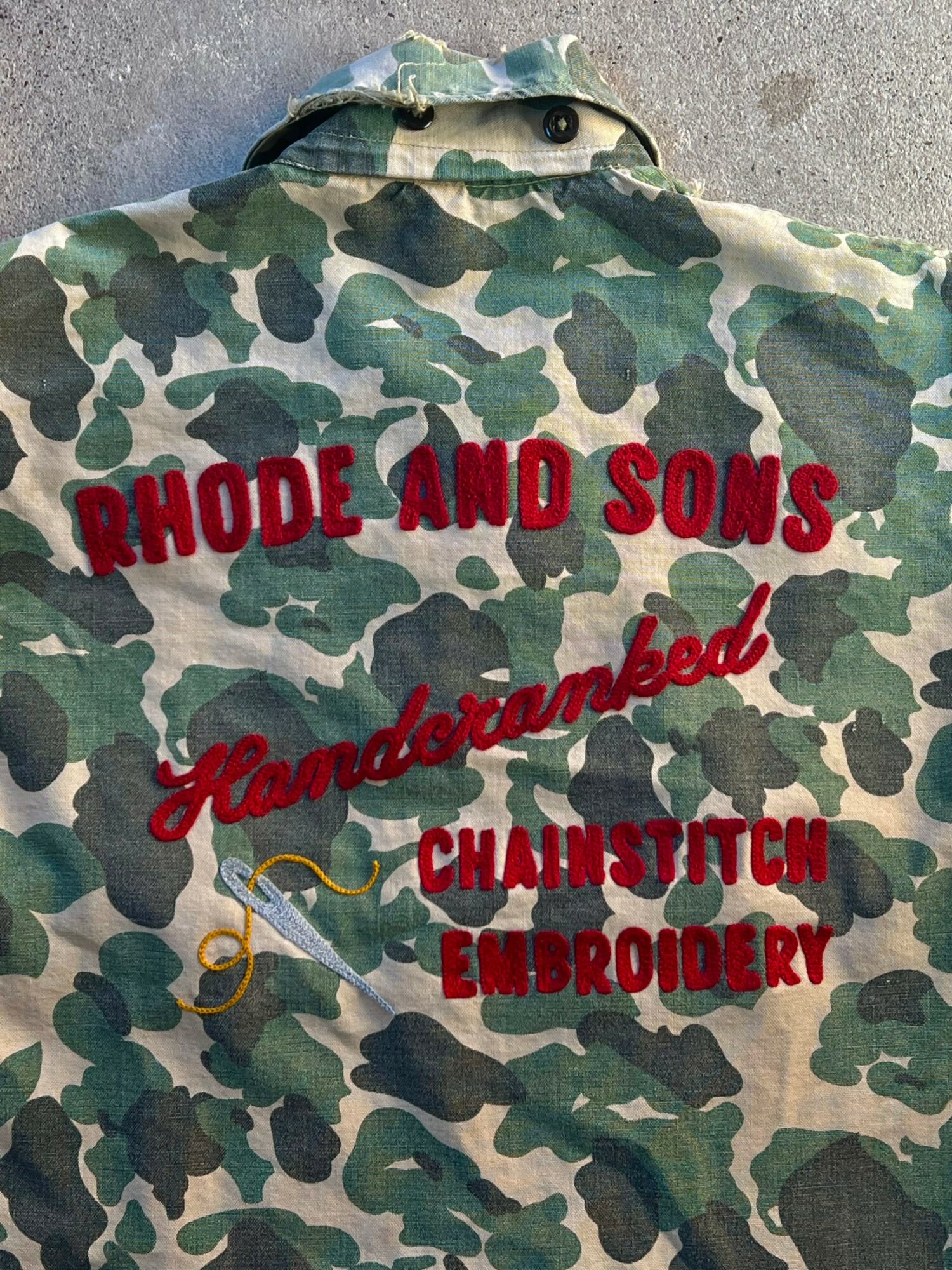 Camouflage jacket with red embroidered text and a yellow needle design, reading 'RHODE AND SONS' and 'Handcrafted Chainstitch Embroidery'.