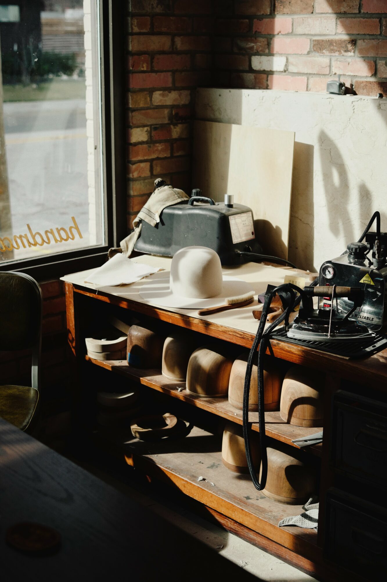 Sunlight illuminates a cluttered wooden shelf with various objects, against a brick wall and window, casting shadows.