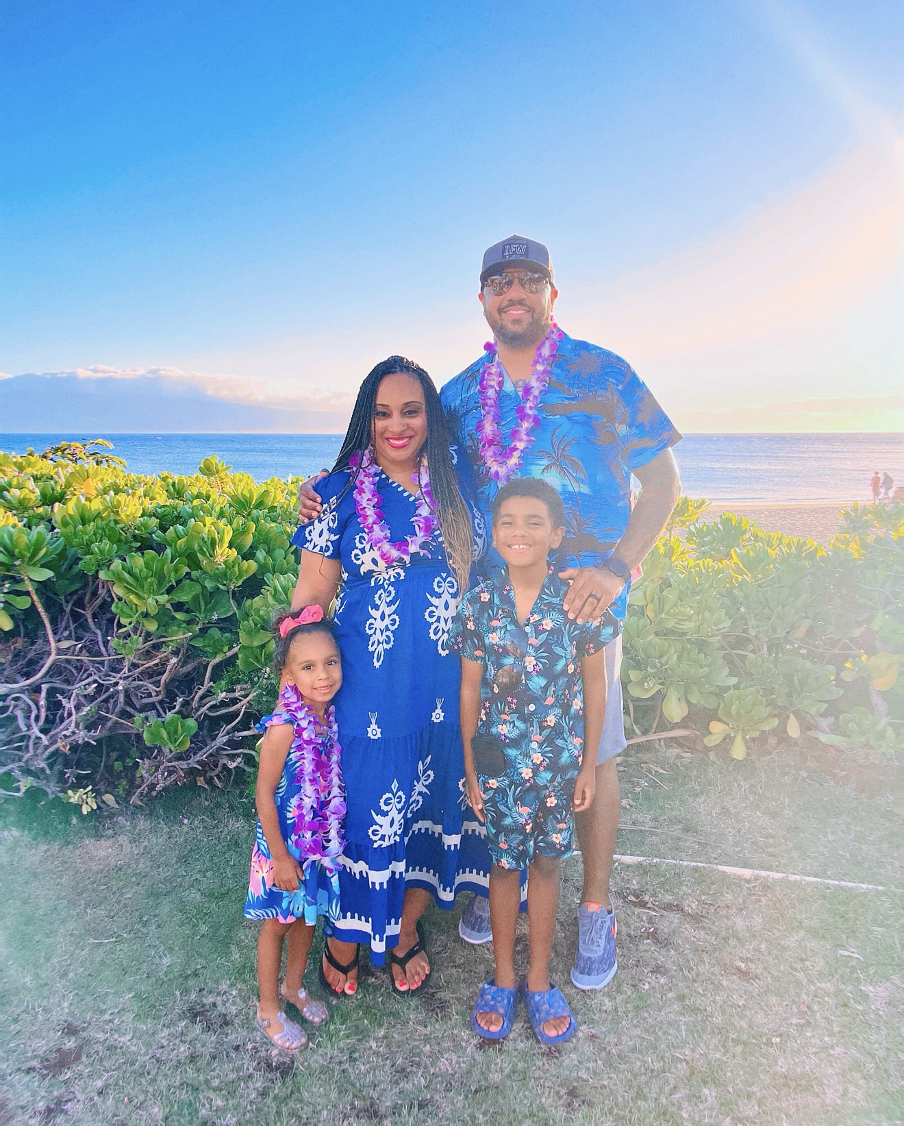 Family of four standing outdoors near water, wearing blue floral clothing, with greenery and sky in background.