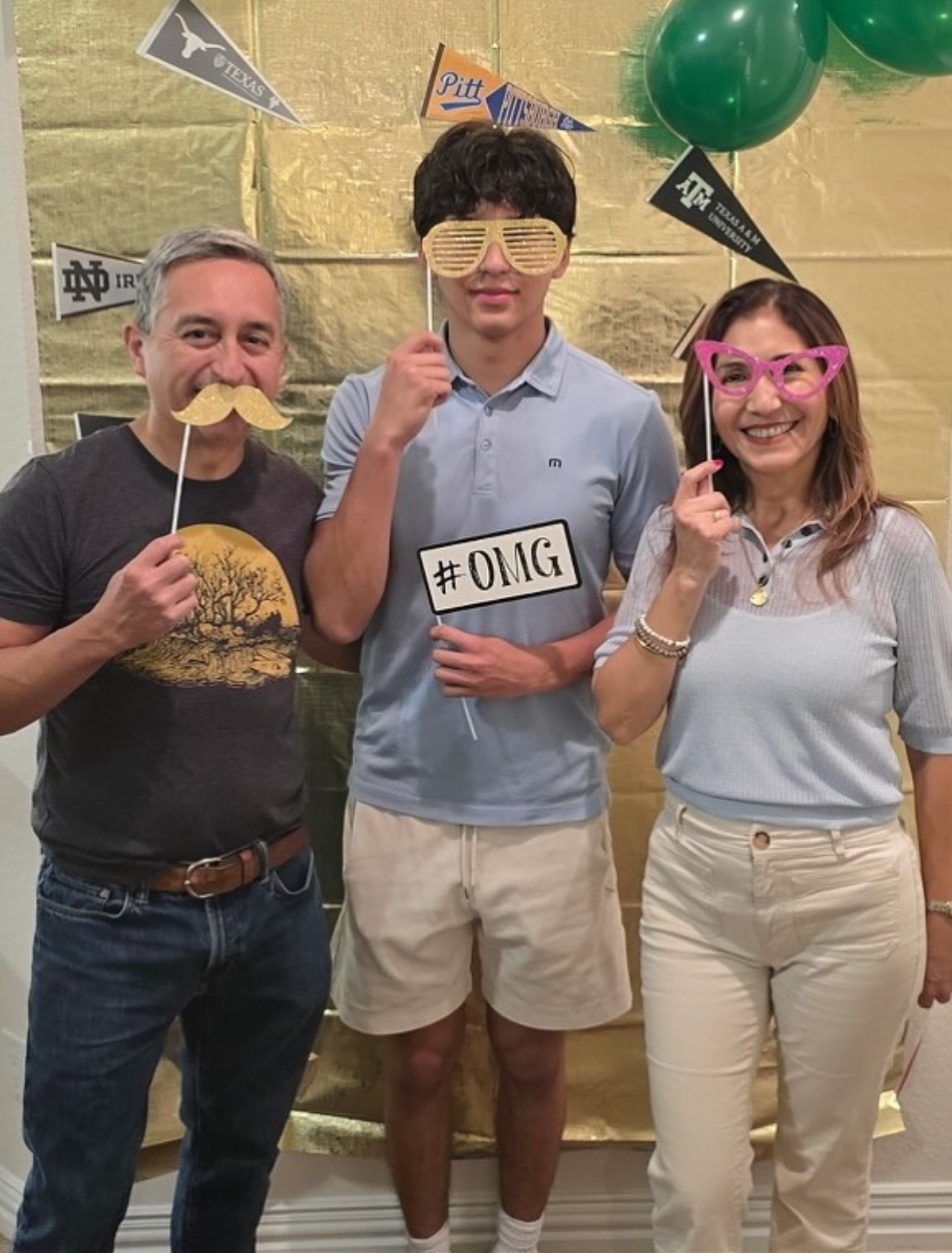 Three people smiling at a celebration, holding fun props and a hashtag sign, with balloons and banners in the background.