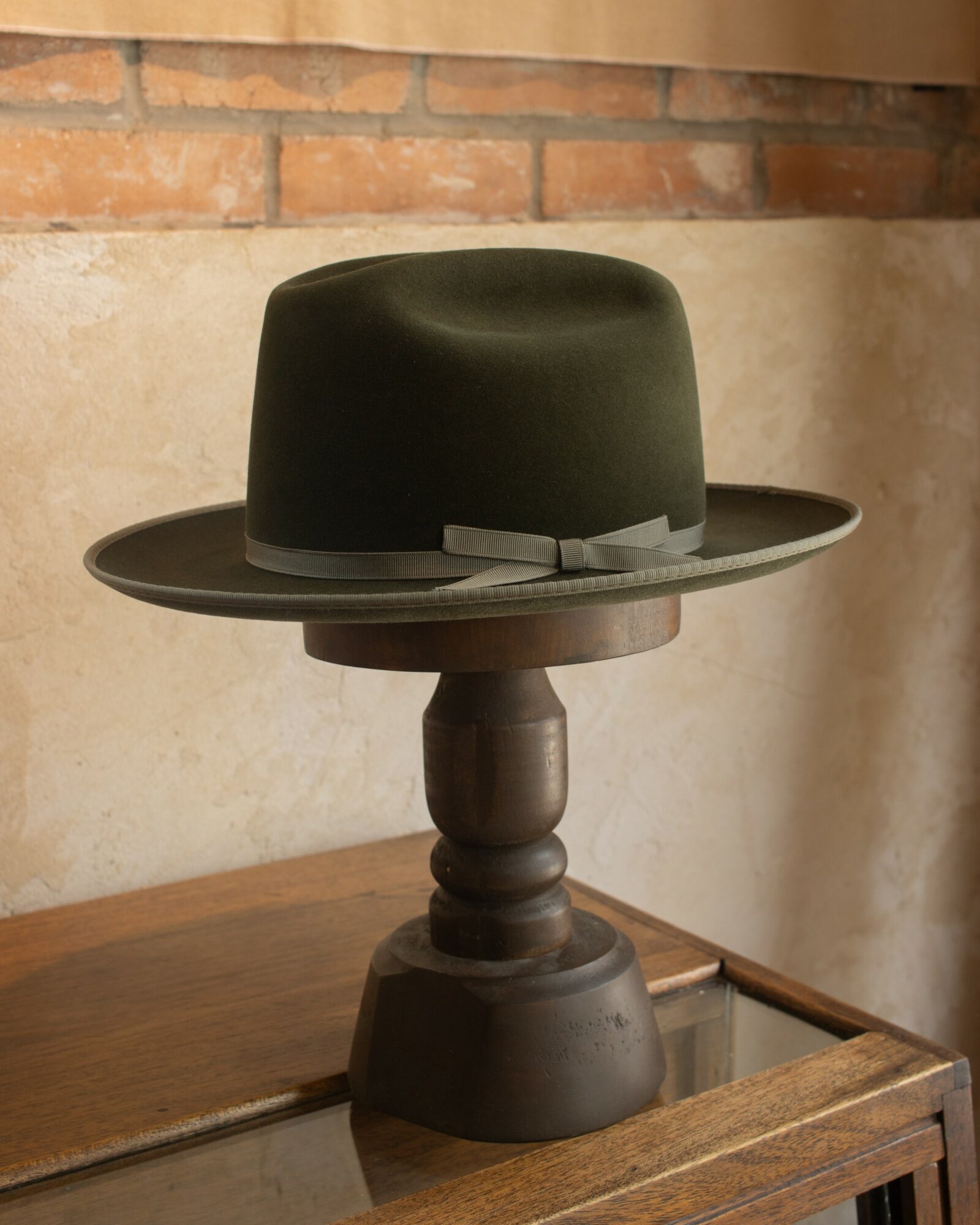 Black hat with a ribbon, placed on a wooden stand, on a wooden table against a brick and plaster wall.