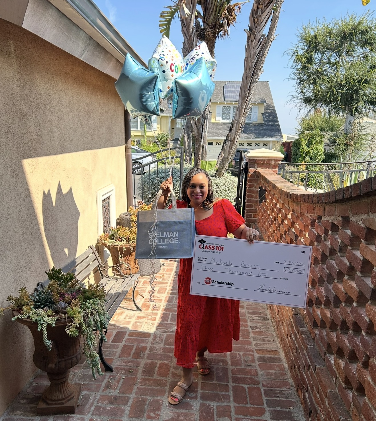 Woman in red dress holding a large check and smiling outdoors on brick pathway, with balloons and plants nearby.