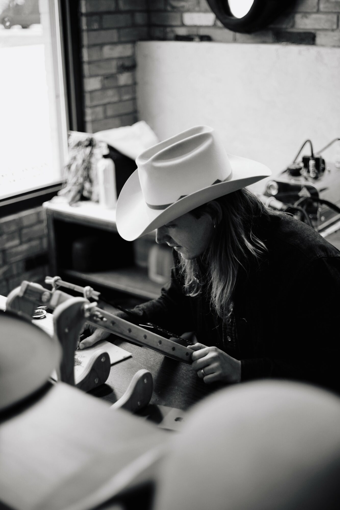 Person wearing a cowboy hat working at a table with various objects, in a room with brick wall and window.