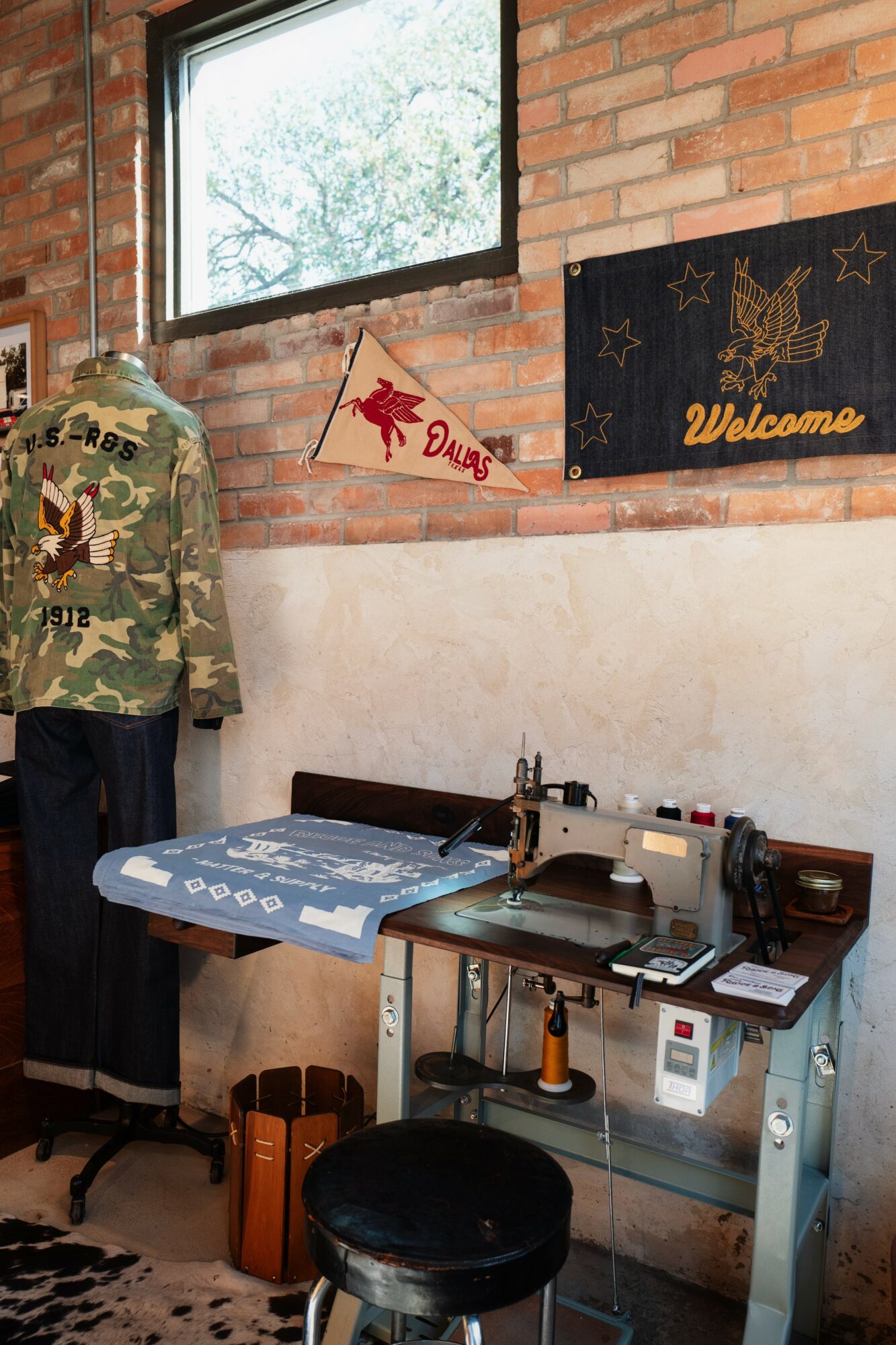 Indoor scene with a sewing machine on a table, a camouflage jacket on a mannequin, and wall decorations, including a welcome sign.