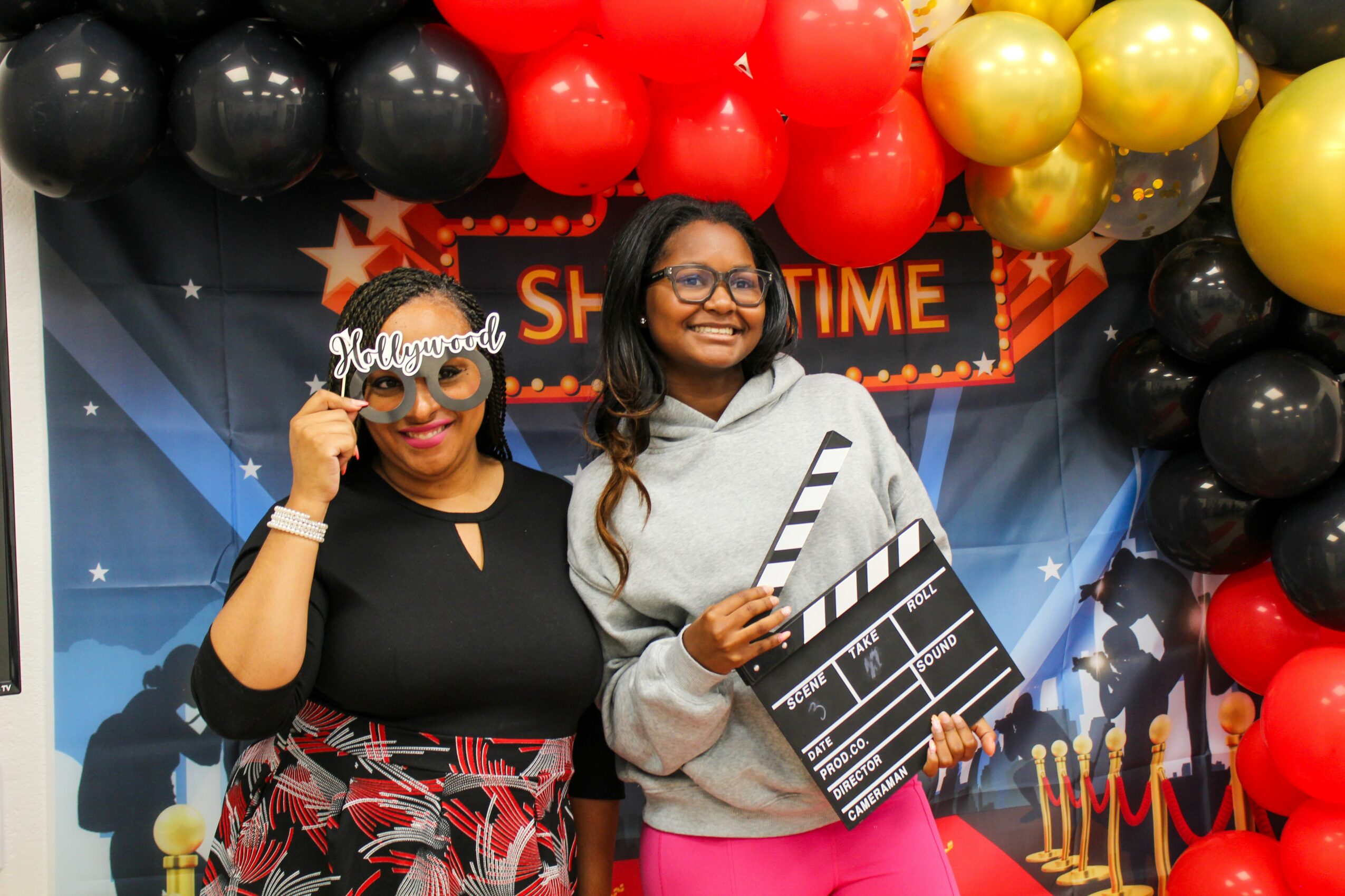 Two women smiling at a celebration with balloons and a movie-themed backdrop, one holding a clapperboard, the other holding glasses.