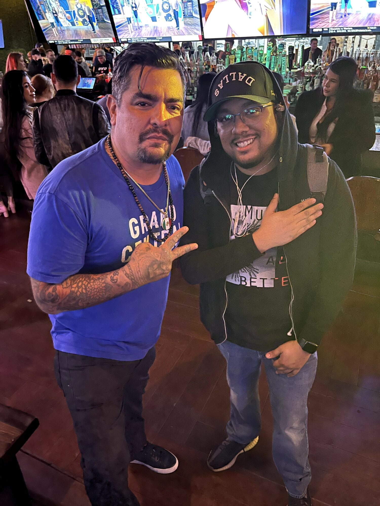 Two men posing together in a crowded indoor setting, one making a peace sign, the other with hand on shoulder, smiling.