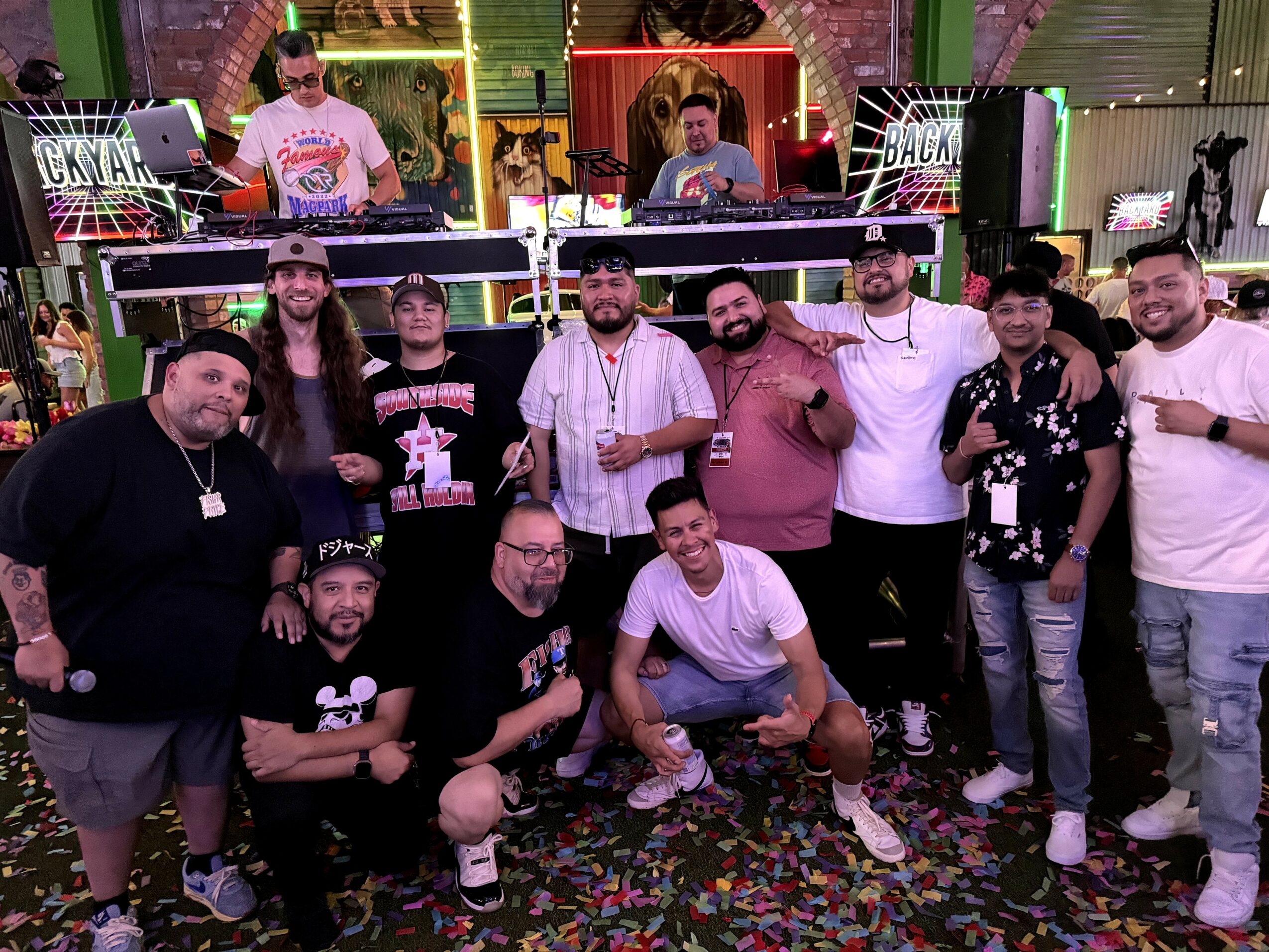 Group of people posing in front of a DJ booth at a party, with colorful lighting and confetti on the floor.