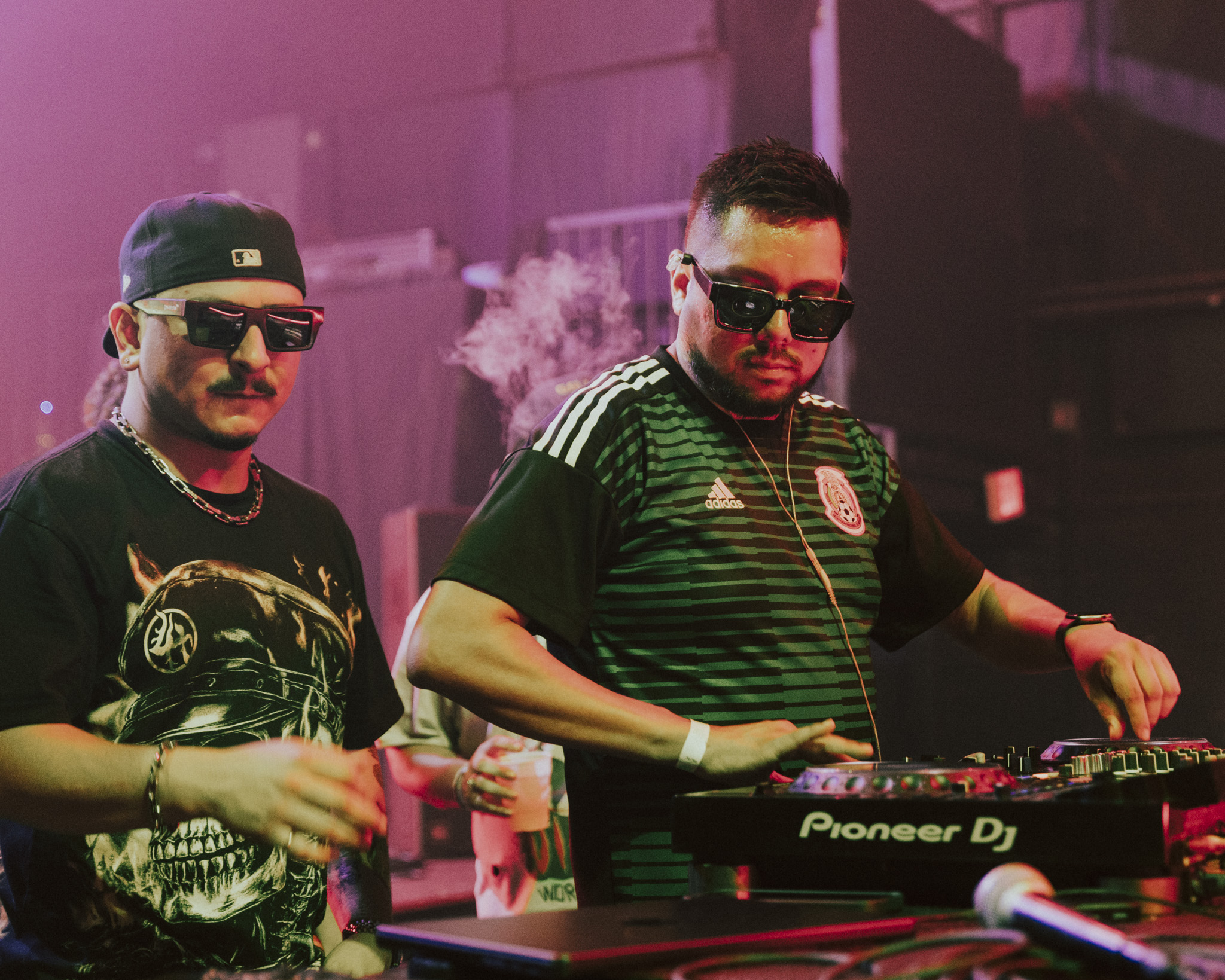 Two DJs wearing sunglasses and headphones perform at a club with pink lighting, one operating a Pioneer DJ controller.
