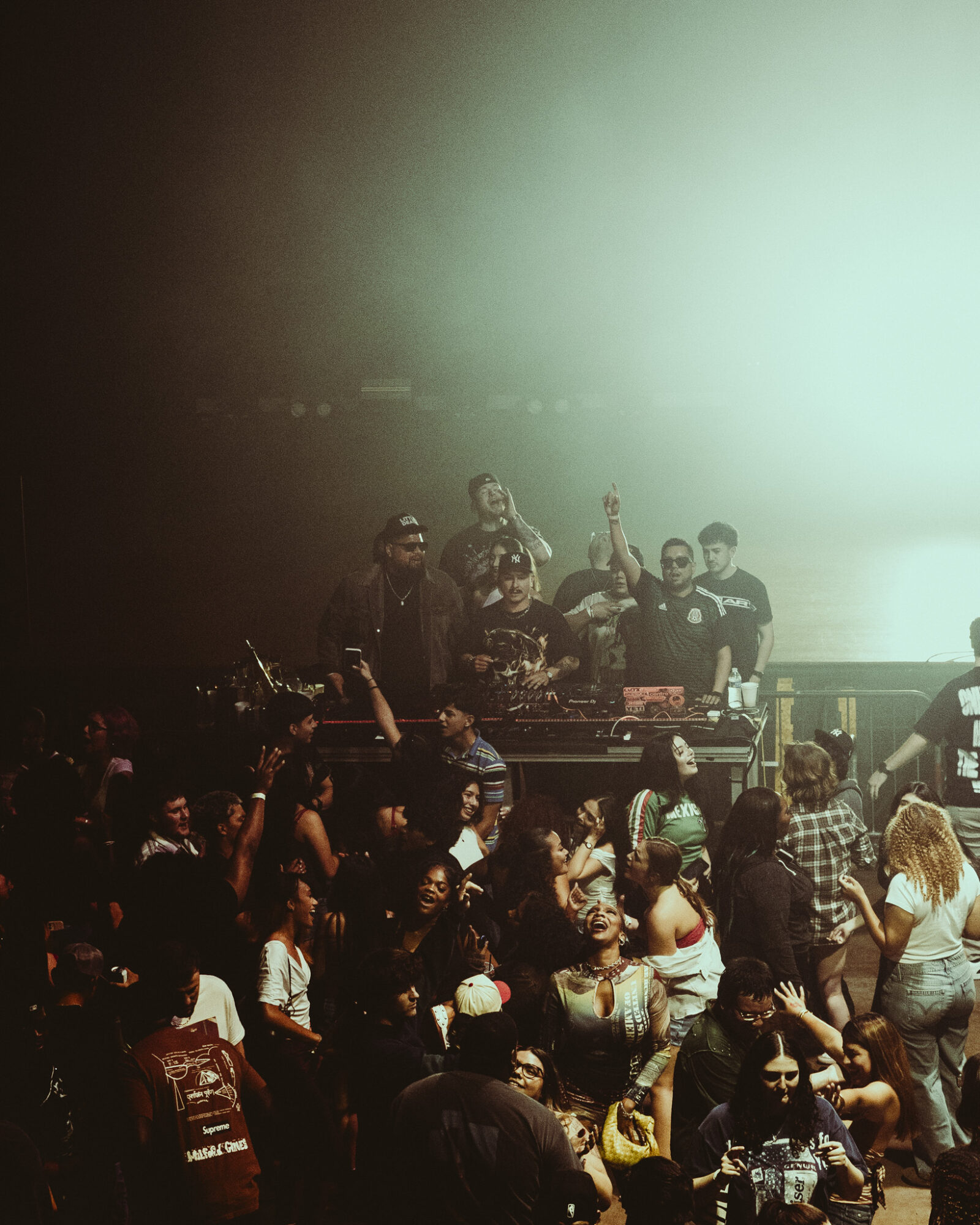 Crowd of people at a concert or event, with some raising hands, in a dimly lit environment with a bright background.