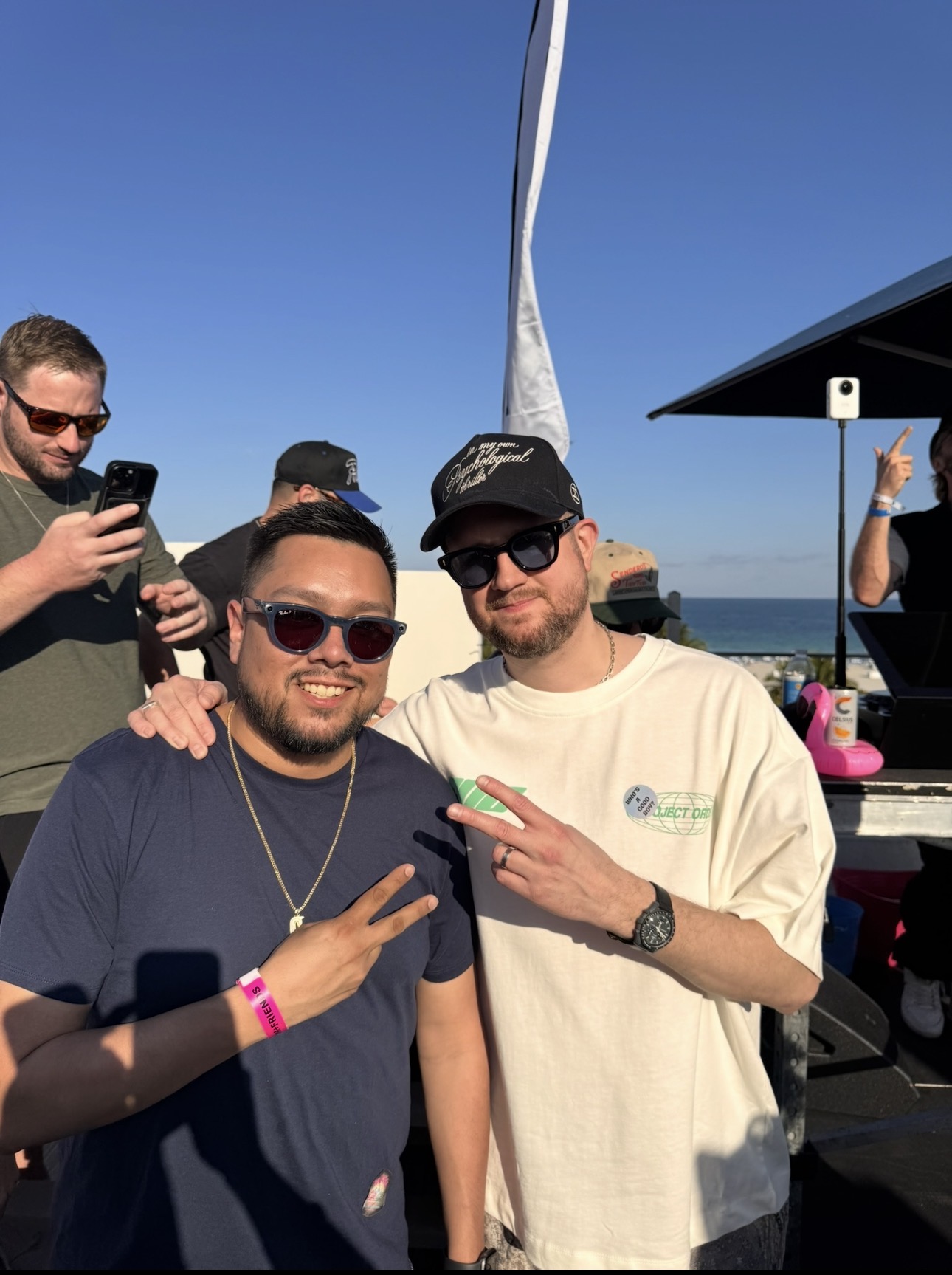 Two men pose outdoors with a clear blue sky, one making a peace sign, the other holding a phone, in a casual setting.