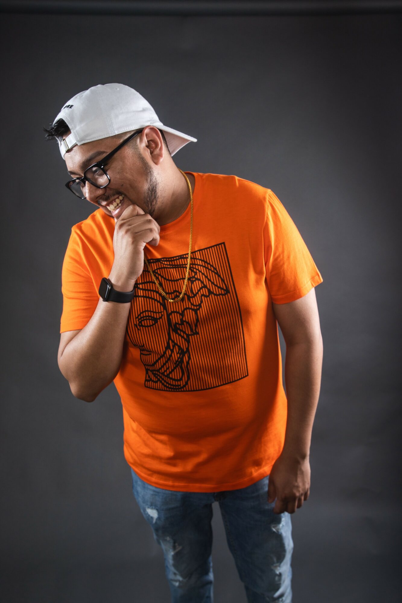 Young man with glasses, white cap backward, orange graphic t-shirt, and jeans, smiling and looking down, against dark background.