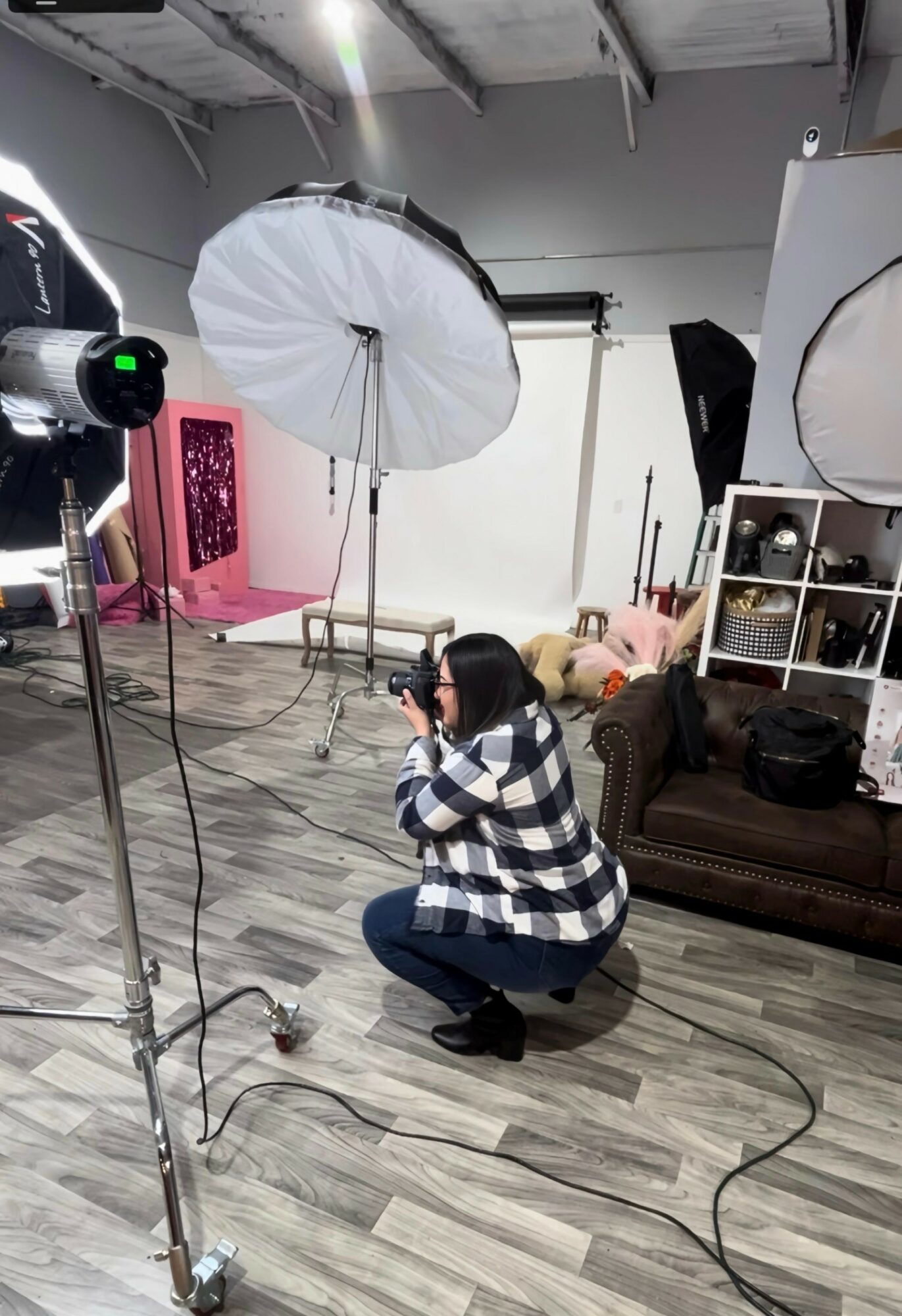 Person crouching in a photography studio with lighting equipment and backdrop, holding a camera, surrounded by studio props and furniture.