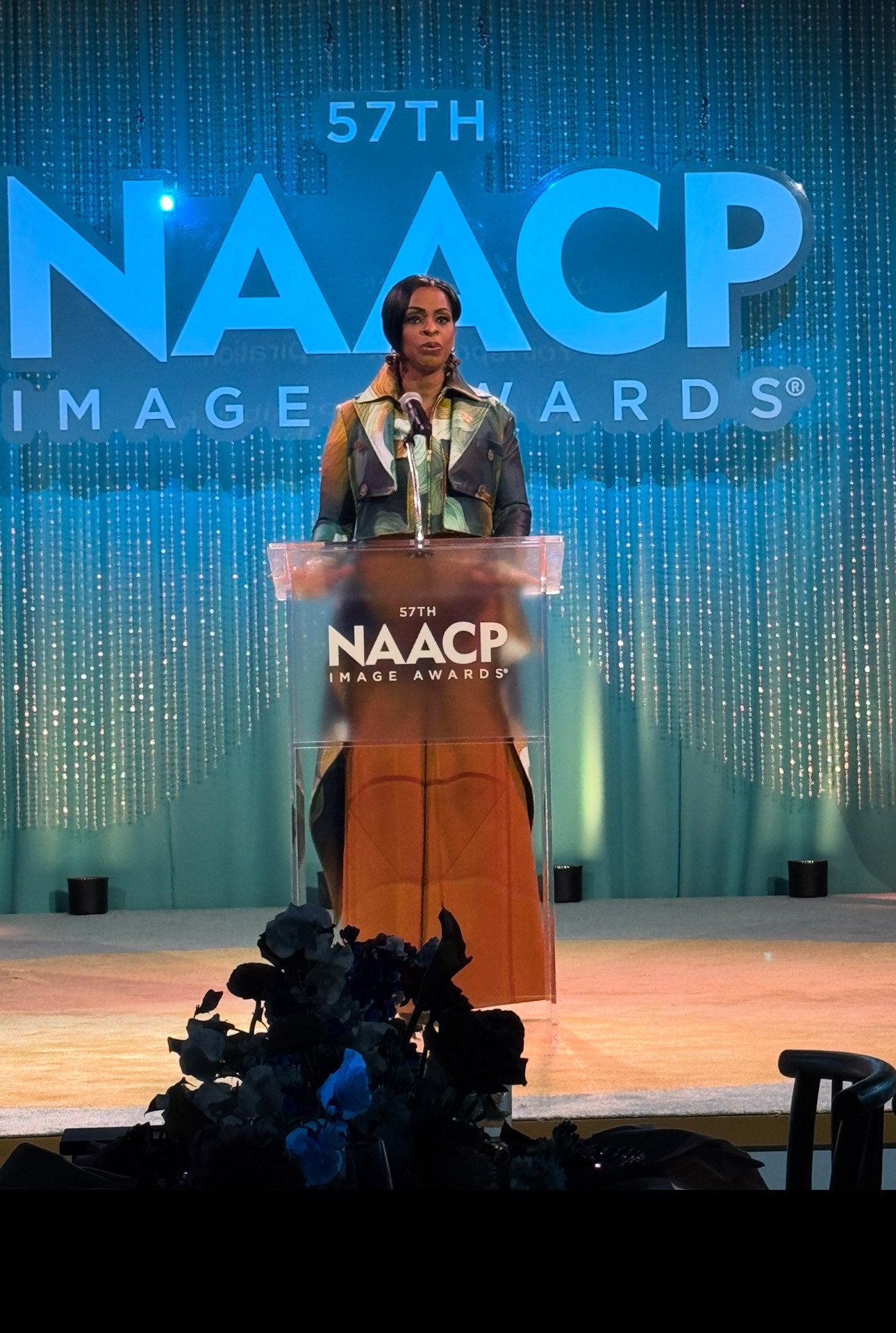 Woman speaking at podium during NAACP Image Awards, stage with large blue NAACP logo in background.