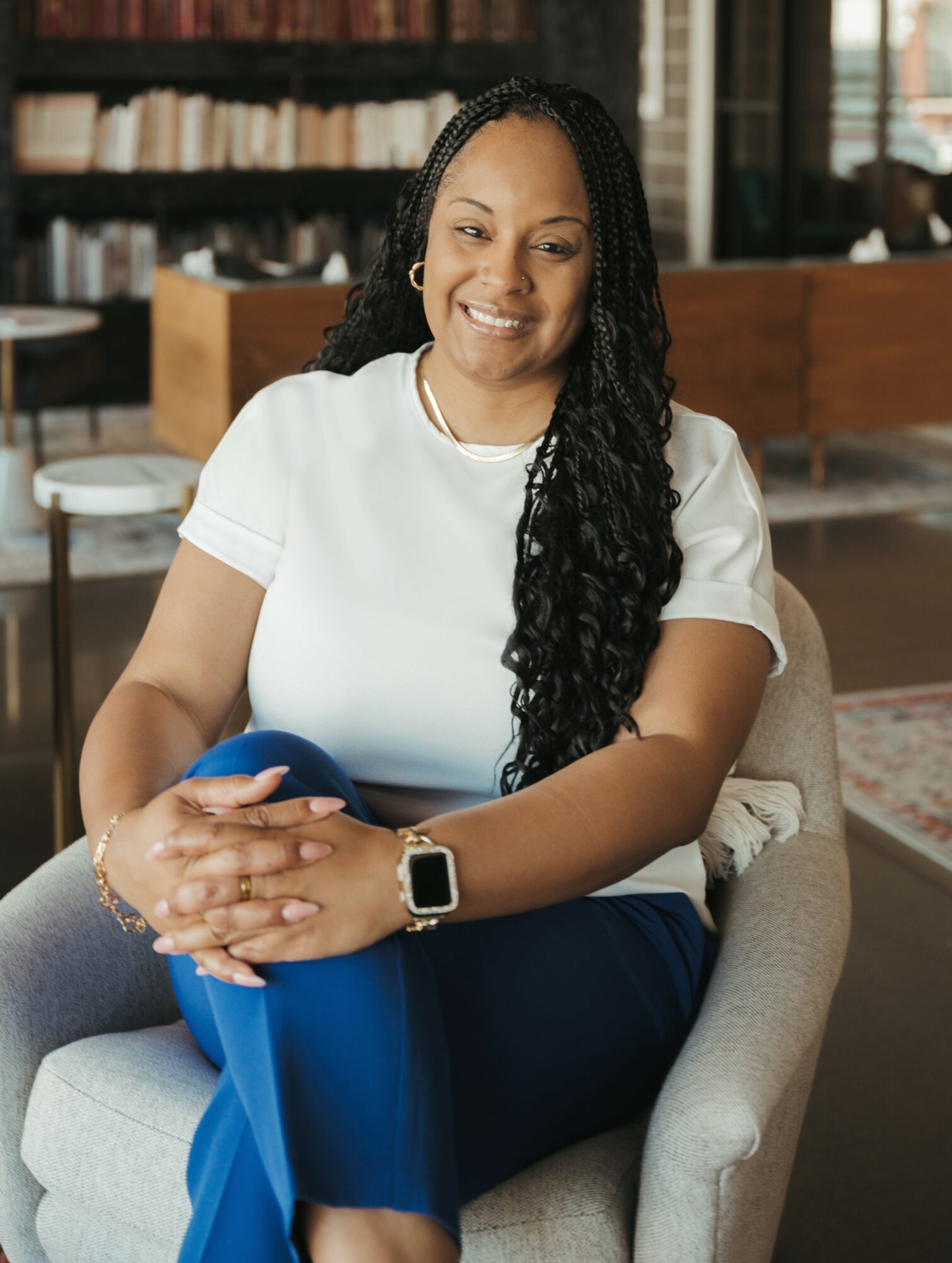 Woman with long curly hair smiling, sitting in a chair in a library or bookstore, holding a phone, wearing a white shirt and blue pants.