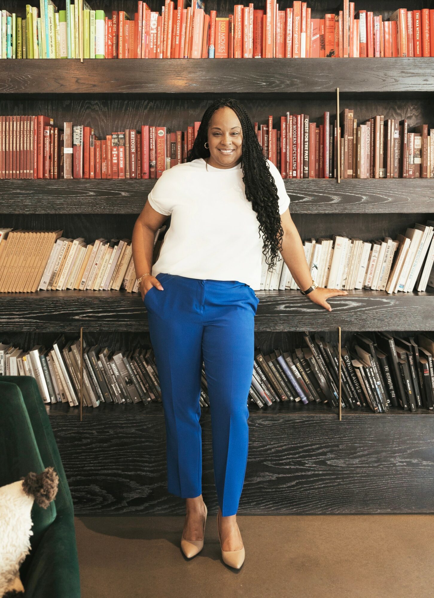 Woman standing in front of a bookshelf with colorful books, smiling, wearing a white top and blue pants, beige heels.