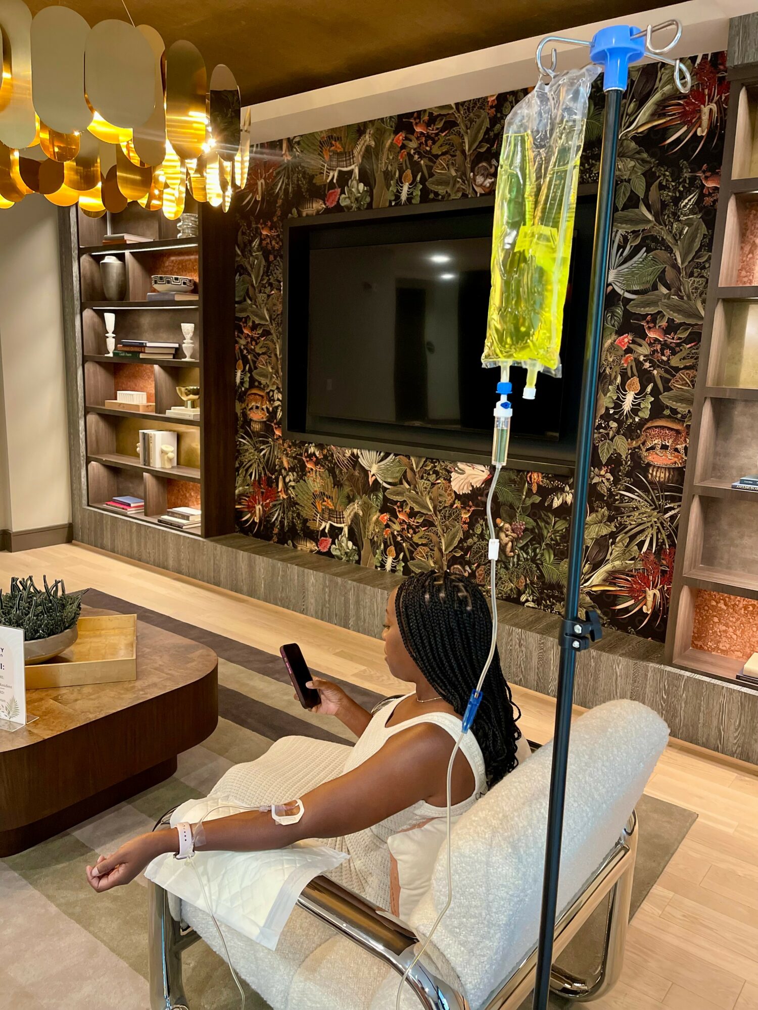 Person lying on a hospital bed with IV drip, in a room with a floral wall, TV, and shelves with dishes.