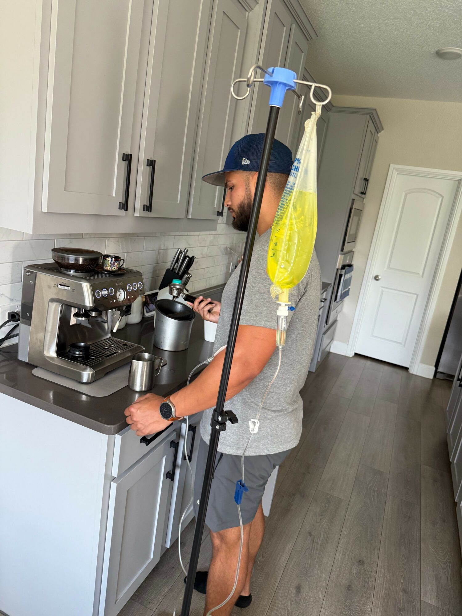 Person in a kitchen preparing a drink with a blender, wearing a gray tank top and shorts, with a yellow bag hanging from a stand.