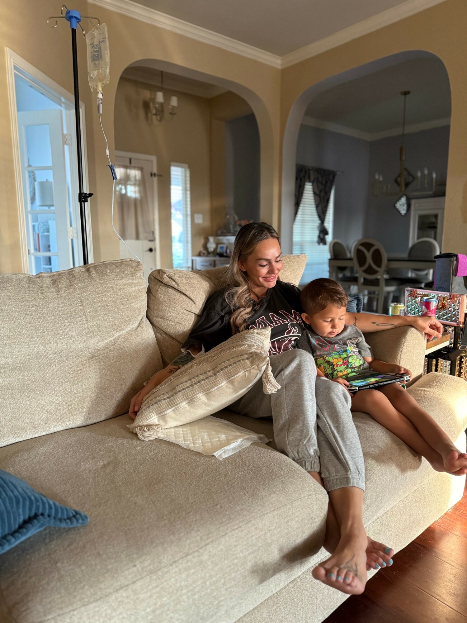 Woman and child sitting on a beige sofa in a living room, smiling and playing with a tablet, with pillows around them.