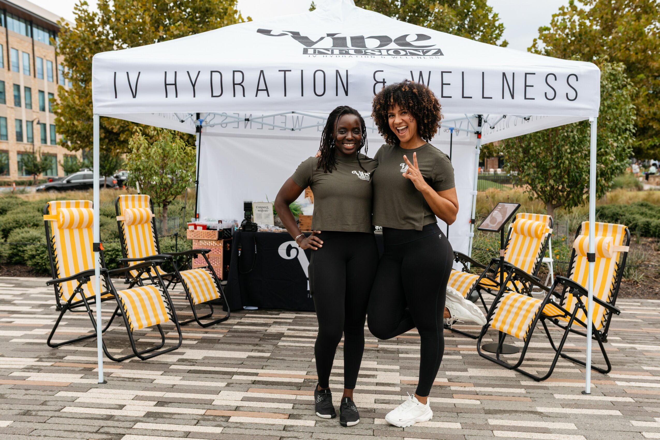 Two women smiling and posing in front of a hydration and wellness tent outdoors, with chairs and trees in the background.