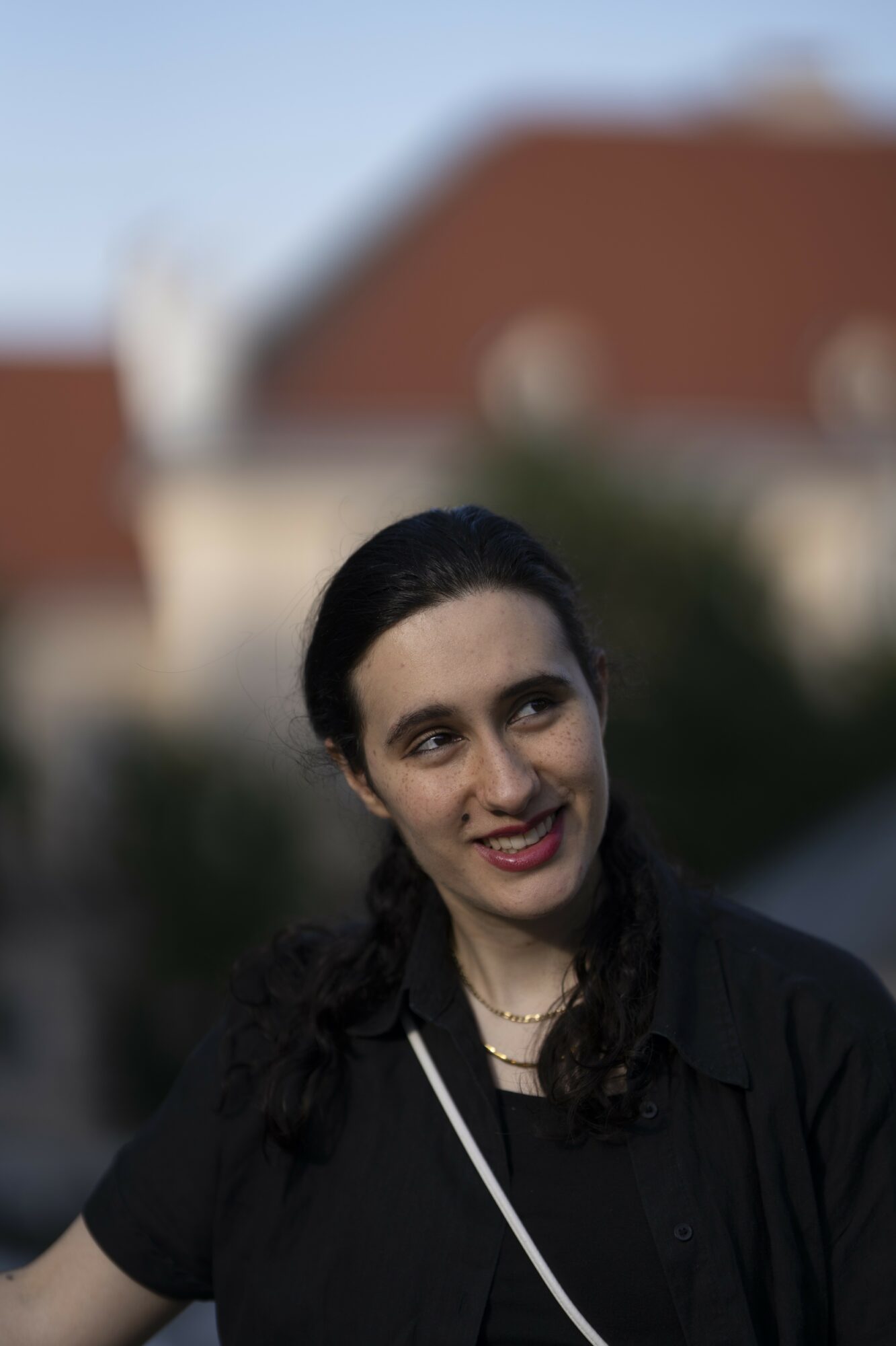 Young woman with dark hair smiling outdoors, wearing a black top and necklace, with buildings in the background.