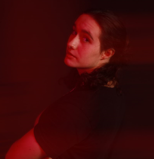 Young person with long hair looking over shoulder in dark setting with red lighting, facing right.