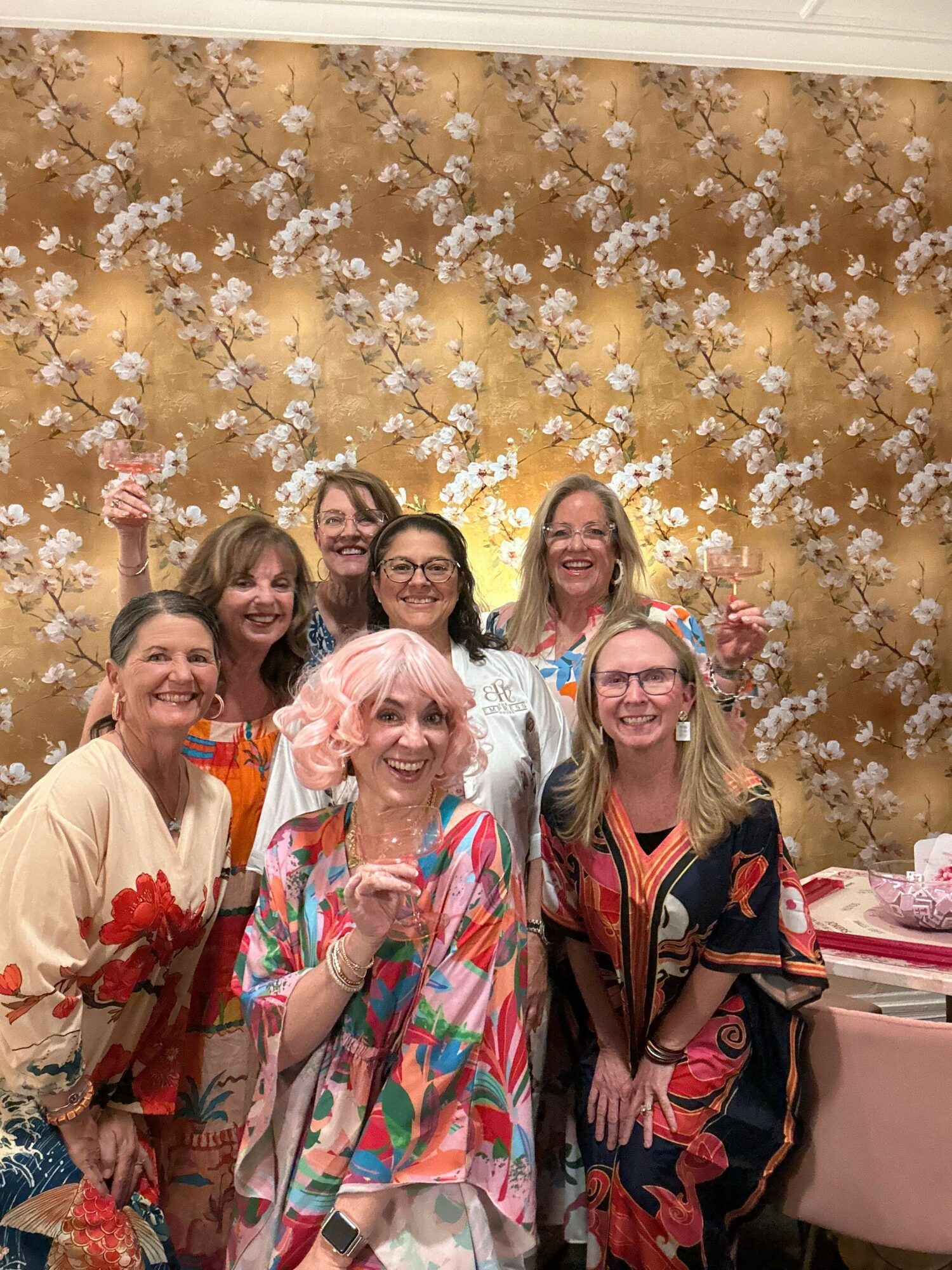 Group of six women smiling, standing in front of a floral backdrop with white flowers and gold background.