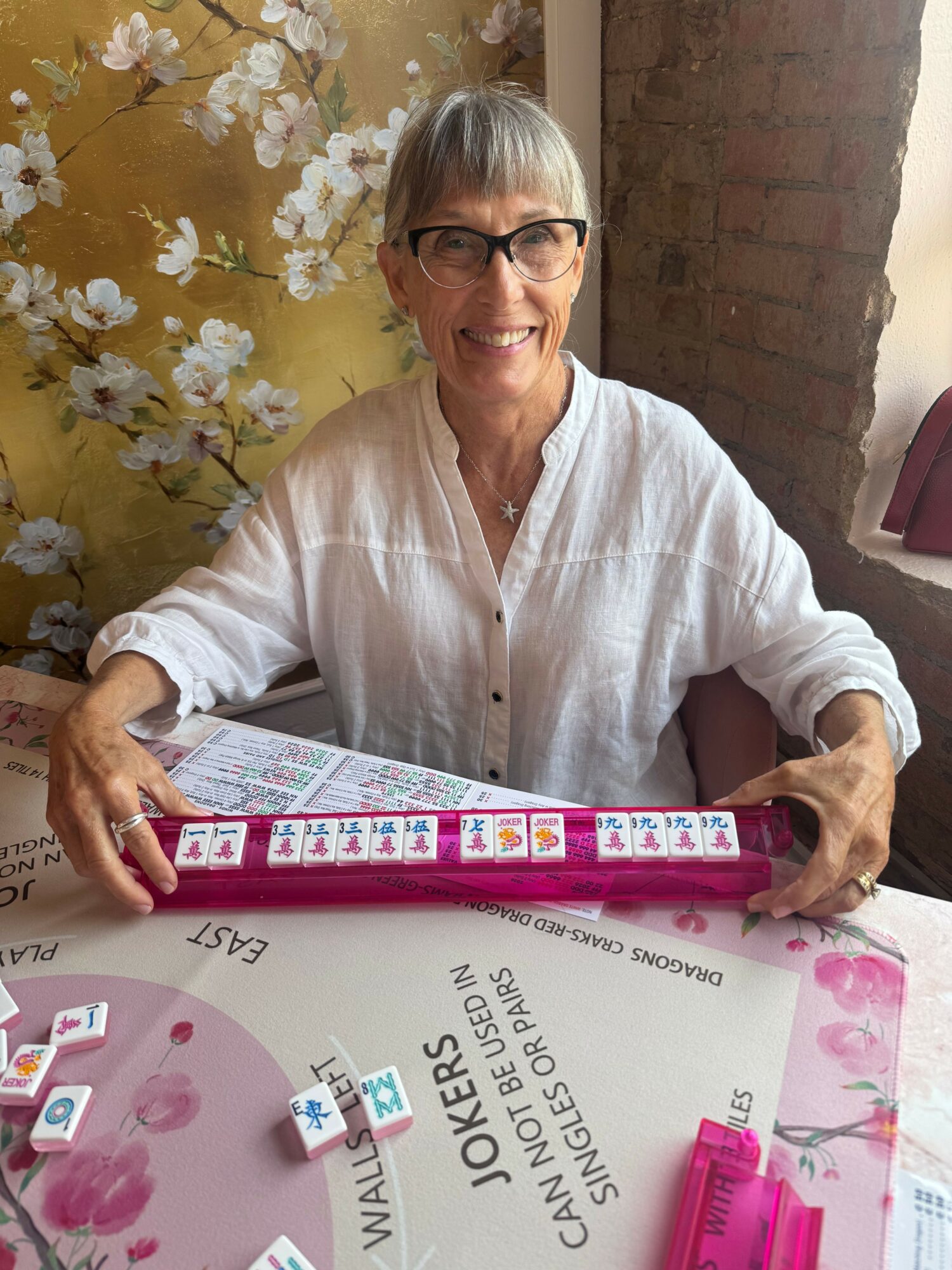 Older woman with glasses and gray hair, wearing a white shirt, sitting at a table with pink and white items, holding a box.