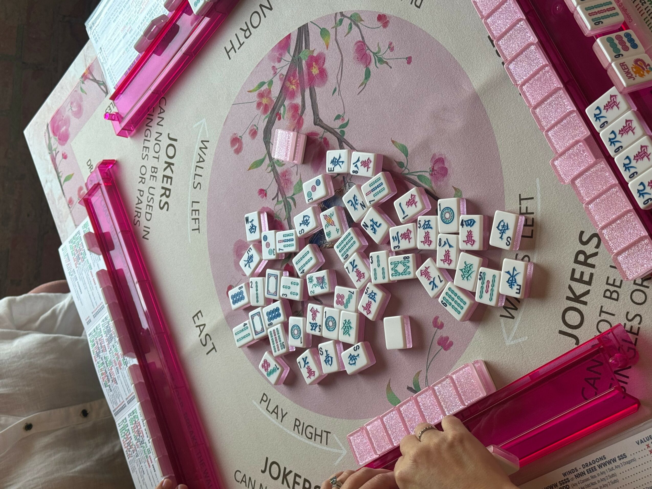 Pink and white Mahjong tiles scattered on a circular game board with pink markers and a hand reaching in.