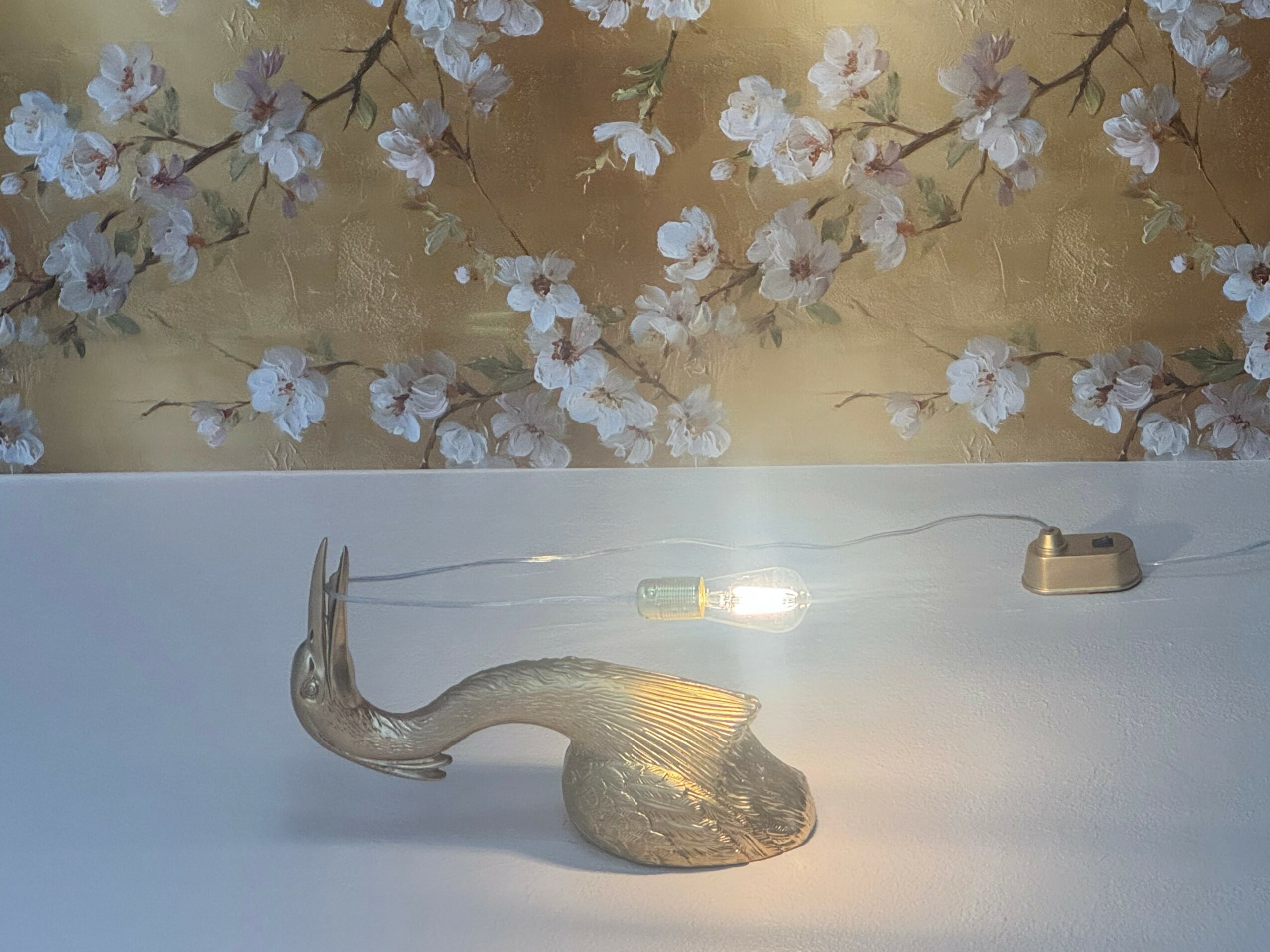 Decorative swan sculpture on a white surface with floral wallpaper background, illuminated by a small light. Word count: 20.