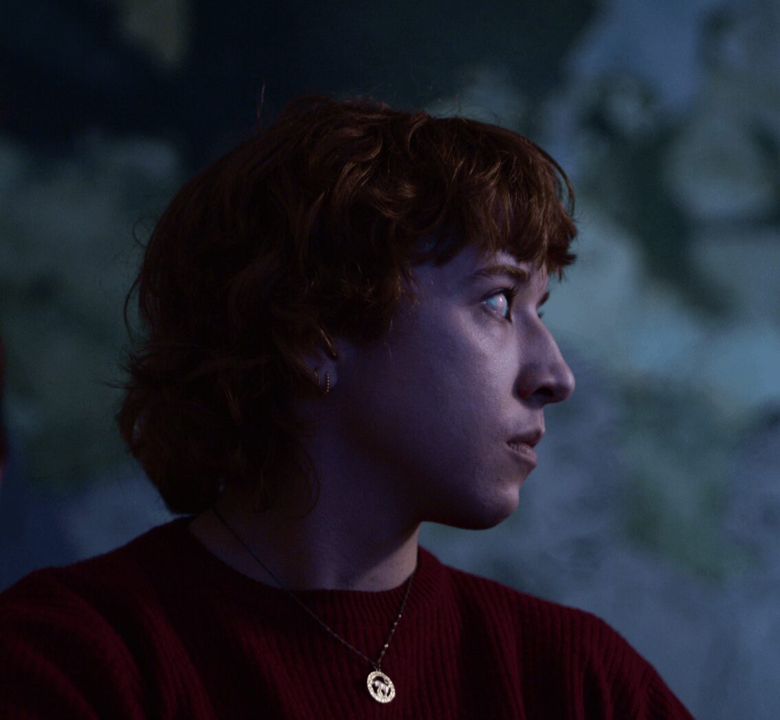 Side profile of a woman with short, curly hair, wearing a red top and a necklace, against a dark background.