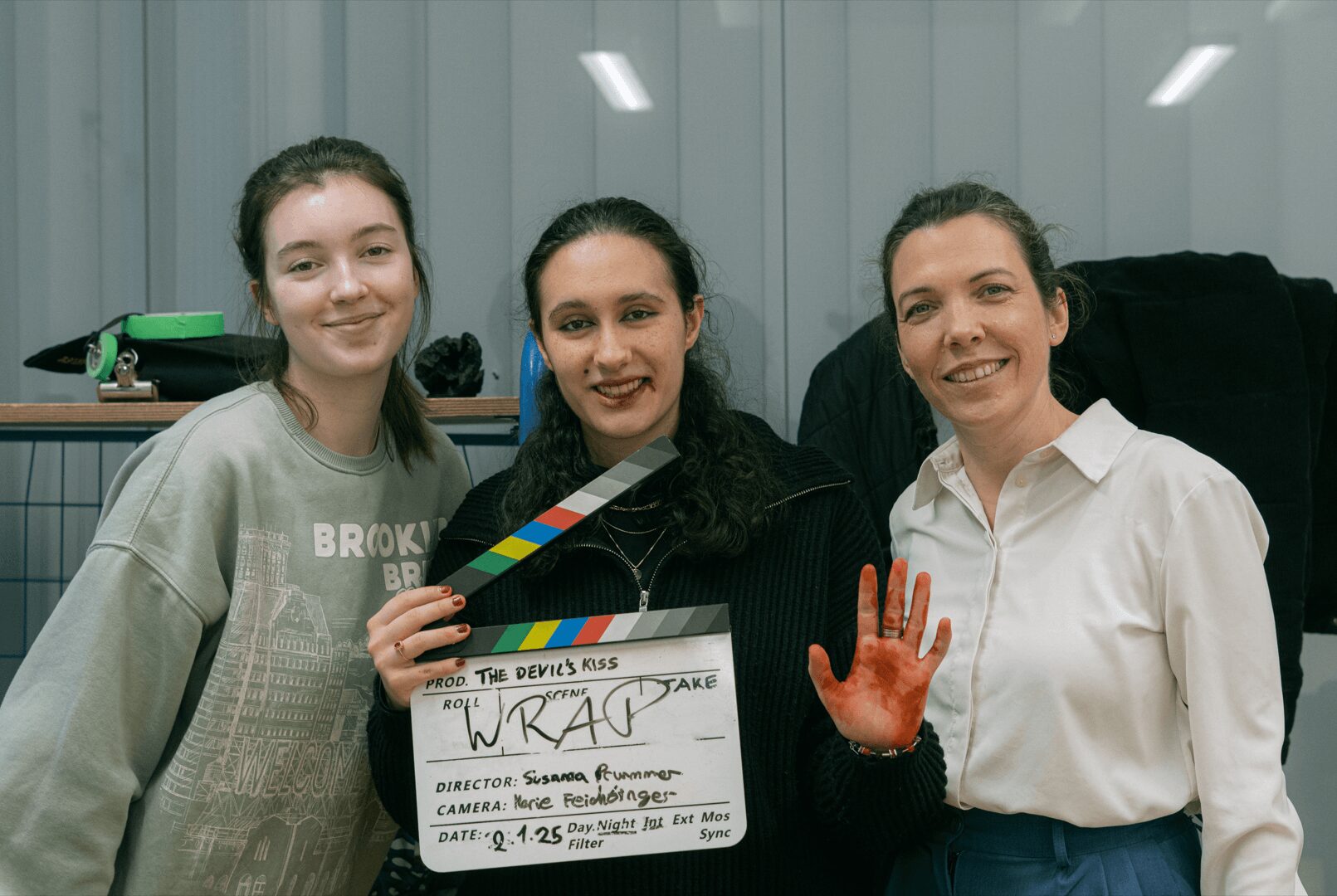 Three women smiling indoors, one holding a clapperboard, another showing a painted hand, in front of a window with vertical blinds.
